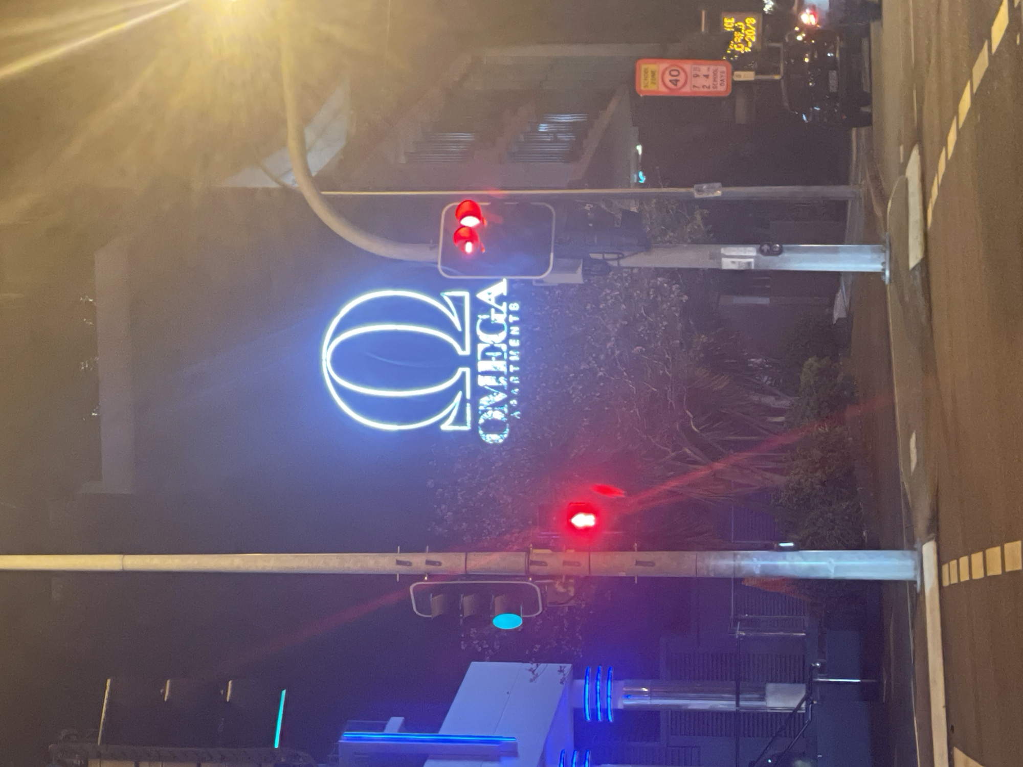 The image depicts a nighttime street scene at an intersection in Brisbane, Australia, specifically near the Omega Apartments. A large, illuminated neon sign for "OMEGA APARTMENTS" is prominently displayed on a building, featuring a stylized Omega symbol in blue and white. A traffic light is visible on the right side of the frame, showing red lights. To the left, another traffic light displays a green light. A speed limit sign for a "SCHOOL ZONE" with "40" and specific hours is also visible. Cars are present on the road, and streetlights and building lights illuminate the surroundings, casting reflections on the wet asphalt. The environment appears urban with some vegetation, possibly trees, visible behind the apartment building.
