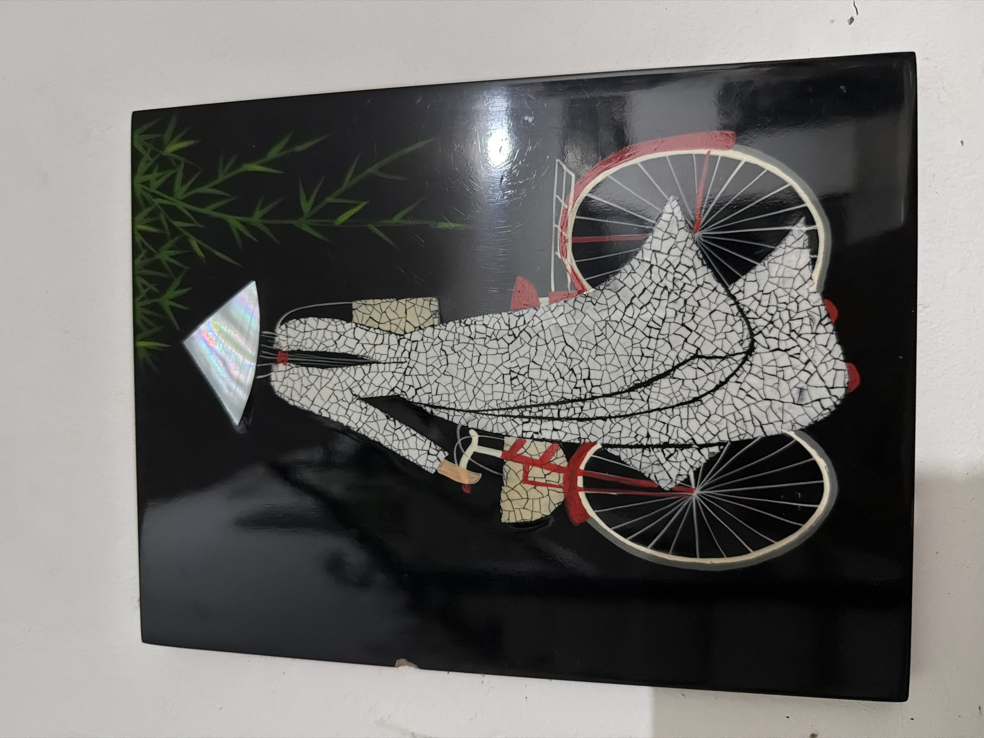 The image displays a black lacquered plaque featuring a mosaic artwork. The artwork depicts a woman in a traditional Vietnamese conical hat and long white áo dài dress, riding a red bicycle. She is carrying a woven bag in her left hand. The background is black, with a few stylized green bamboo leaves in the upper right corner. The mosaic is made of small, irregular pieces of what appears to be mother-of-pearl or a similar iridescent material, creating a cracked-glass effect on the dress and hat. The bicycle has a red frame and spokes, with a light-colored rim. There is a basket on the front of the bicycle. The plaque itself has a glossy finish and shows some minor scratches and smudges. There is no discernible location in Davao City, Philippines depicted.