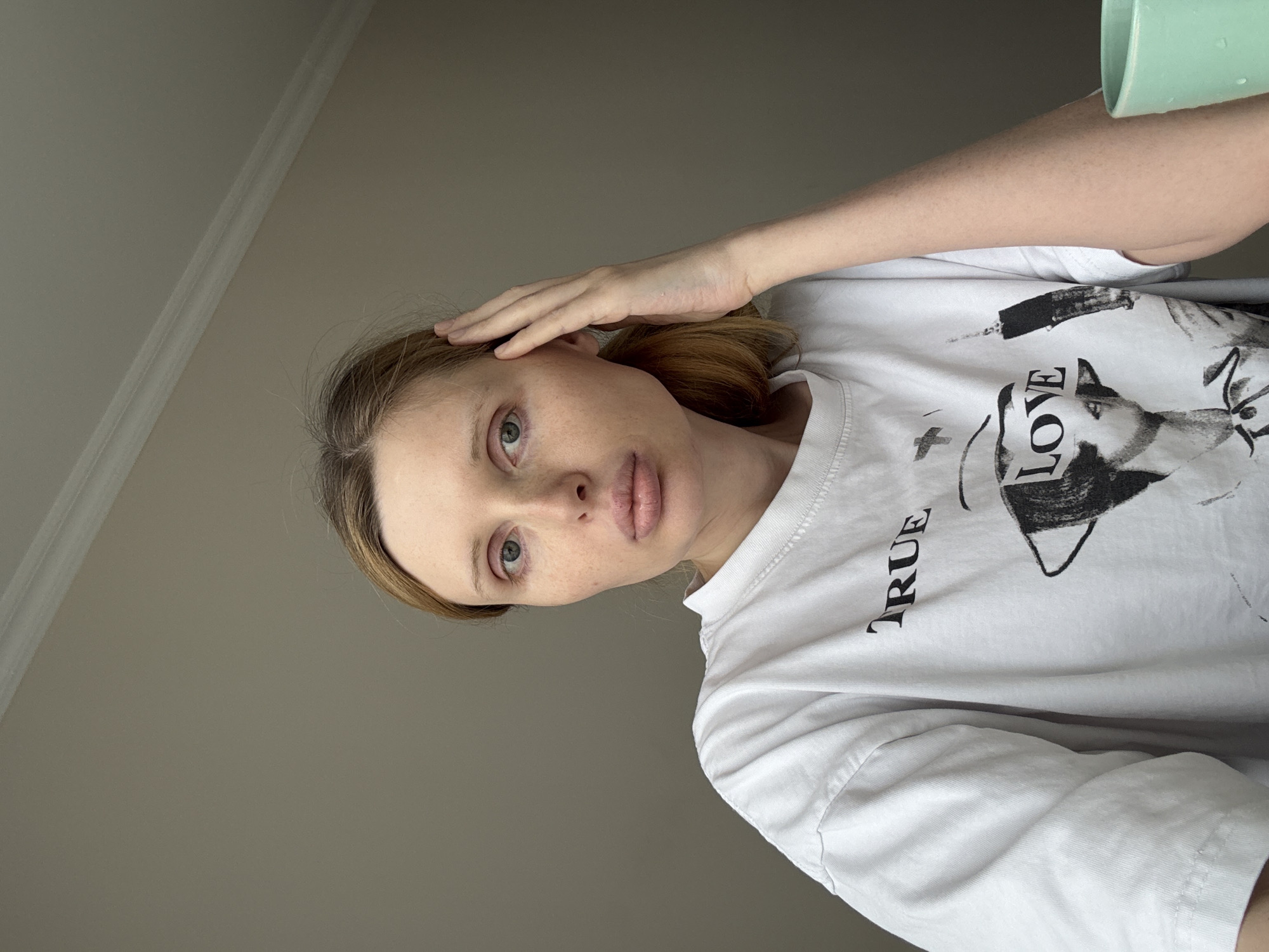 A young woman with light skin and freckles, blonde hair pulled back, and blue eyes looks directly at the camera. She is wearing a white t-shirt with the words "TRUE LOVE" printed on it, along with a graphic image. Her right hand is touching her temple. The background is a plain, light-colored wall with white molding at the top. To the right, a light green mug is partially visible. The scene appears to be indoors, in an unspecified location.