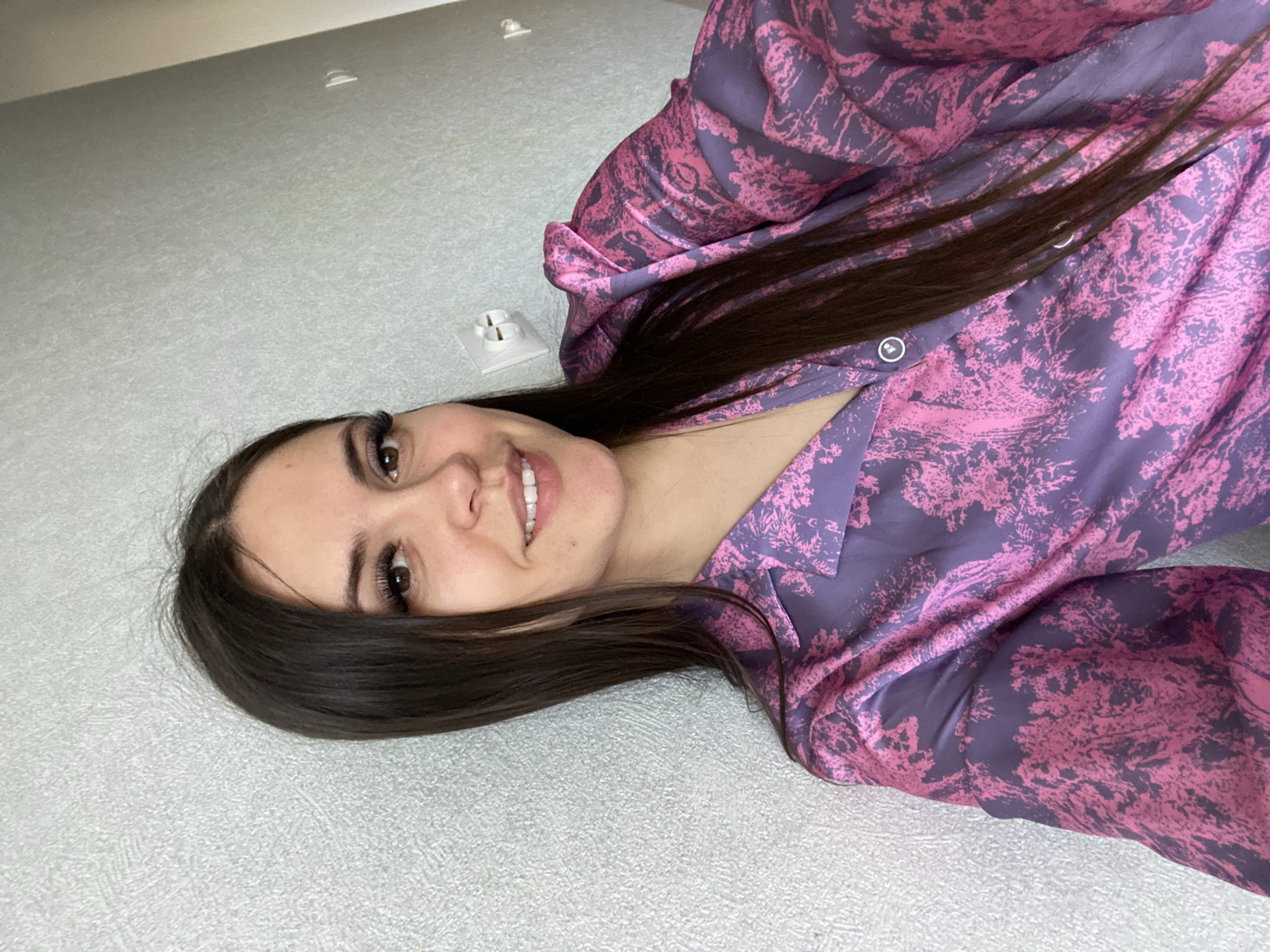 An image shows a young woman in a rotated orientation, appearing to be lying down or leaning. She smiles warmly at the viewer, revealing white teeth. She has long, dark brown hair, brown eyes, and prominently long eyelashes. Her hair spreads across her shoulders and the patterned top she wears.
The top is long-sleeved, featuring an abstract pattern in shades of purple and pink, resembling foliage or trees. Two light-colored buttons are visible near her collarbone.
The background is a light grey, textured surface, likely a wall or floor. Two white electrical outlets are visible on this surface, suggesting an indoor setting. One is partially visible near her hair, and another is further up in the frame. Her expression conveys cheerfulness and relaxation. The activity is posing for a photo indoors. No visible text is present. The location context is an indoor setting in Chaikovsky, Russia.