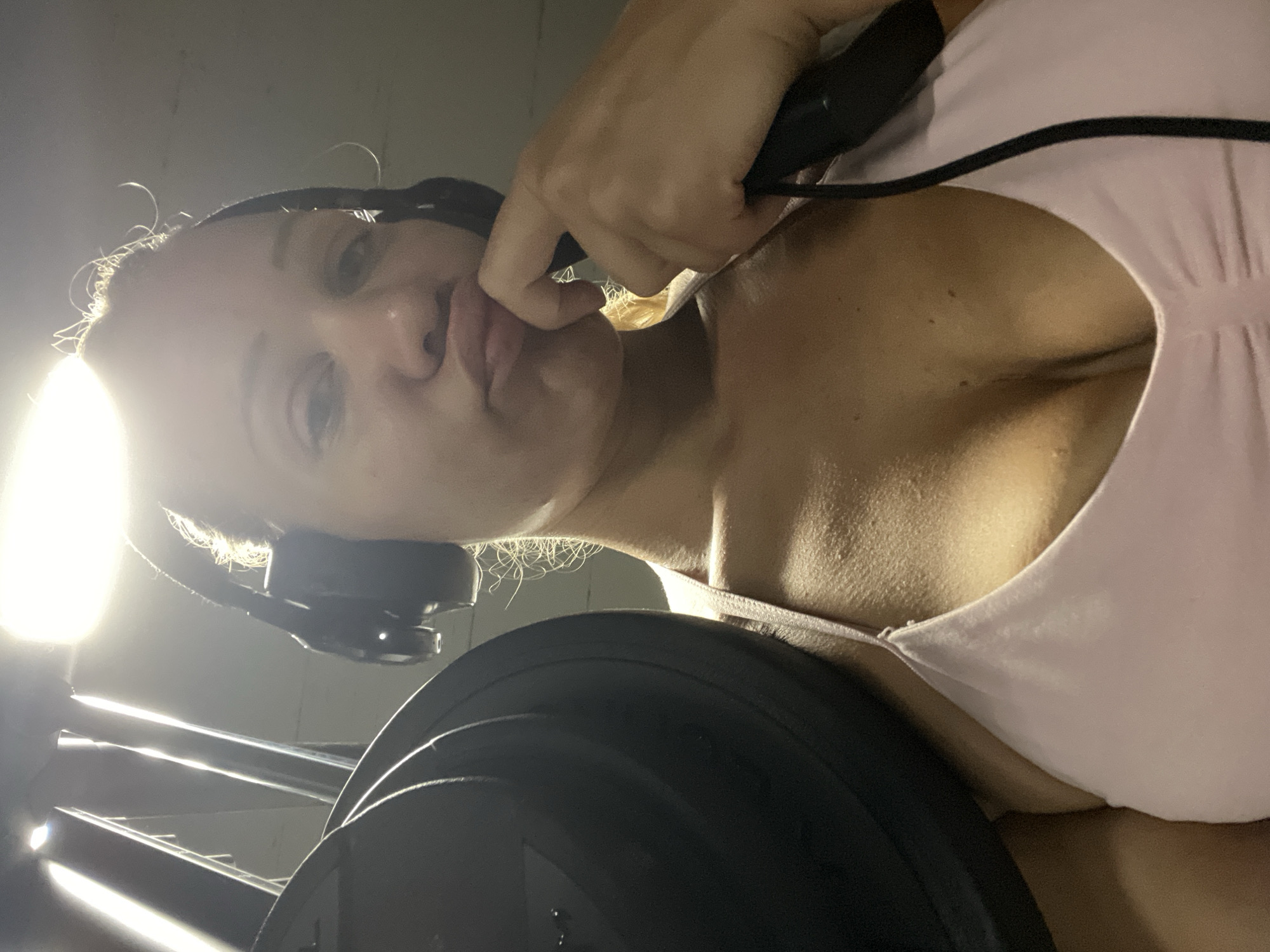The image shows a close-up, from a low angle, of a woman in a gym. She is wearing headphones and a light pink sports bra. A weight plate is visible on her left shoulder. Her right hand is touching her chin, and she is looking at the camera with her lips pursed. The lighting is bright and overhead. The image does not contain any information about the city of Altötting, Germany.
