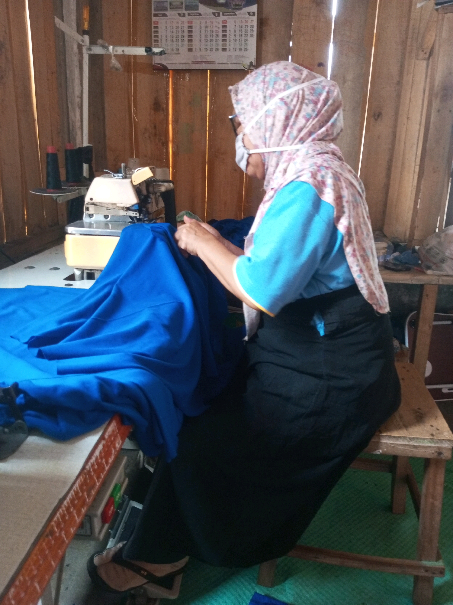 In Kalibeber, Indonesia, a person wearing a headscarf, glasses, and a face mask is sewing on a white sewing machine. The person is dressed in a blue shirt and a black skirt. The sewing machine is on a white table with a large piece of blue fabric draped over it. In the background, there are wooden walls and a calendar on the wall. There are also two spools of black thread on a stand next to the sewing machine. A wooden stool is beside the table.
