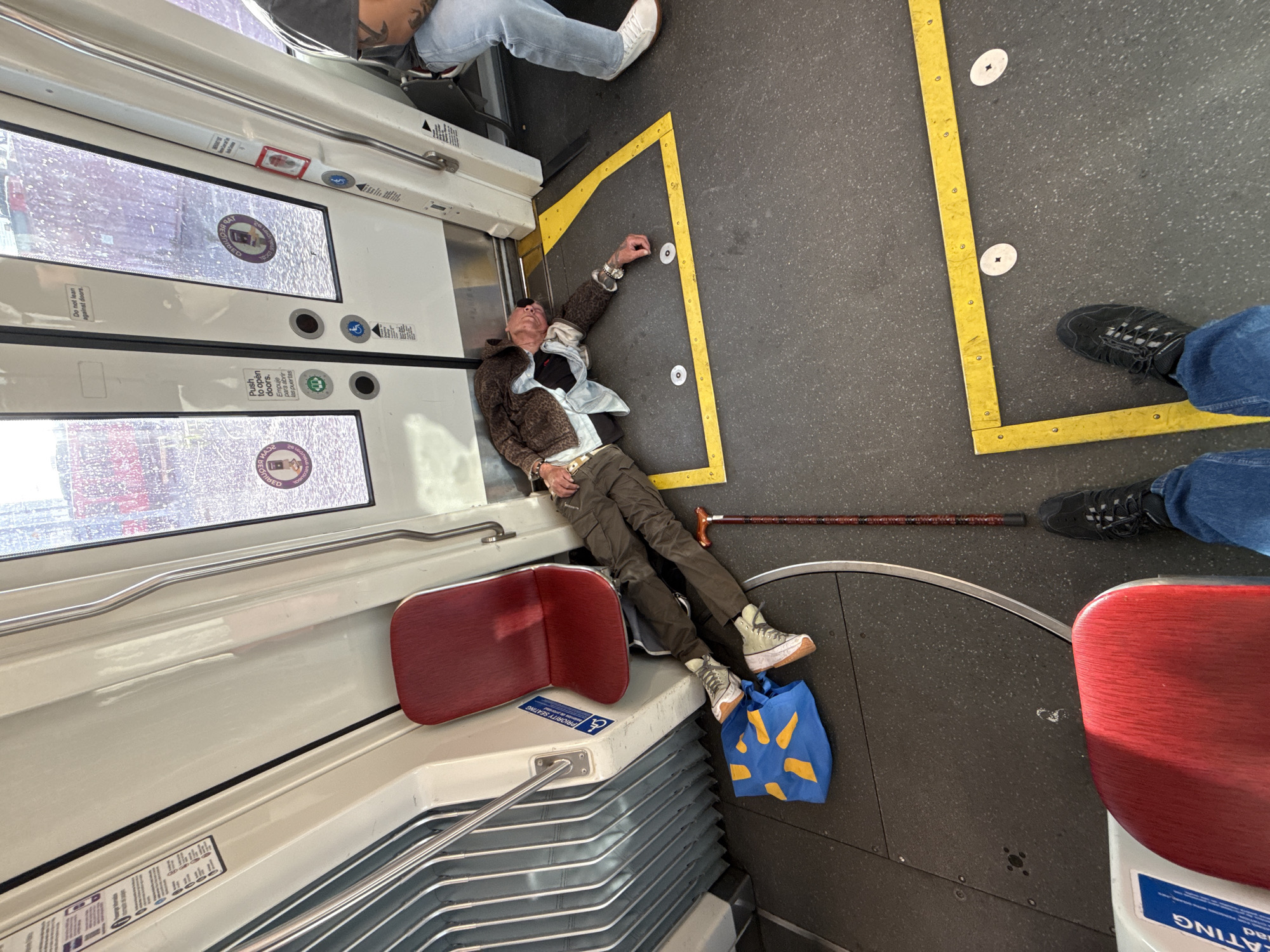 A man appears to be unconscious or asleep on the floor of a train car in San Diego, California. He is lying on his back with his head against the wall near the doors. He is wearing a brown patterned jacket over a light blue shirt and khaki pants. A cane lies on the floor next to him, and a blue shopping bag with a yellow design is at his feet. Another passenger is visible in the background, sitting on a seat. The train car has red seats and a gray floor with yellow markings. The doors appear to be automatic.
