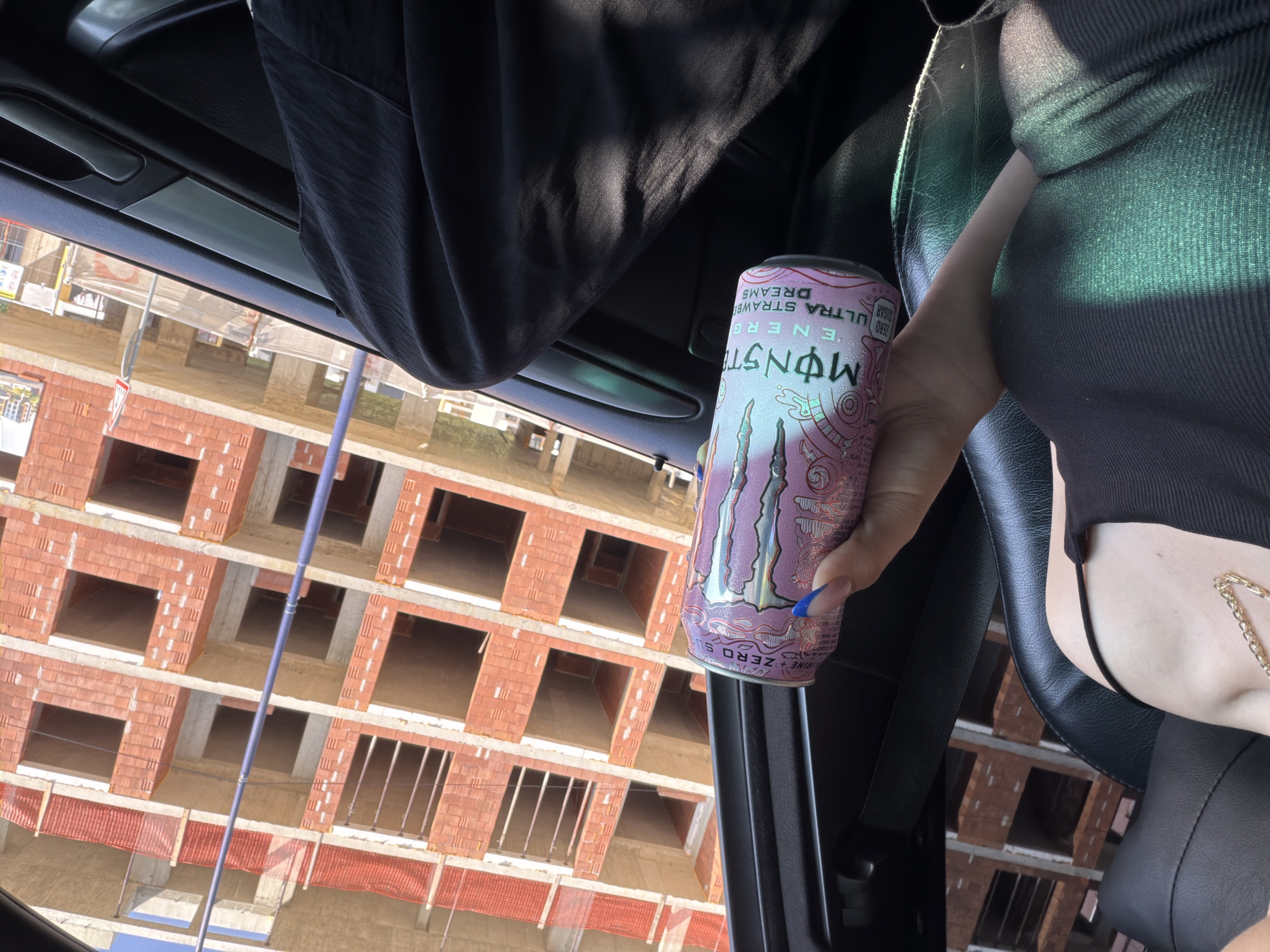 The image captures a partial view of a person inside a vehicle, looking out at a construction site in Pazardzhik, Bulgaria, during daytime. A person, whose arm, hand with blue-painted nails, and part of their torso are visible on the right, is holding a pink Monster Energy drink can. They are wearing a dark, ribbed top, and a gold chain hangs near their shoulder. The car interior features dark plastic and fabric elements, with sunlight casting patterns on the seat and person.
The Monster can's label clearly displays "MONSTER ENERGY ULTRA STRAWBERRY DREAMS ZERO SUGAR" and "200" (likely caffeine content). Other text like "CAFFEINE + ZERO S" and smaller details are also present.
Outside the vehicle, a multi-story building under construction dominates the background. It features exposed red brick (likely ceramic block) walls and concrete floor slabs, with numerous rectangular openings for windows or doors. Orange safety netting is visible on lower levels of the construction. A blue pipe or pole extends vertically across the frame, in front of the building. In the distance, another building and some trees are faintly visible. A red and white yield road sign is also discernible, along with other indistinct signs near the construction site. The scene is brightly lit by sunlight, indicating clear weather.