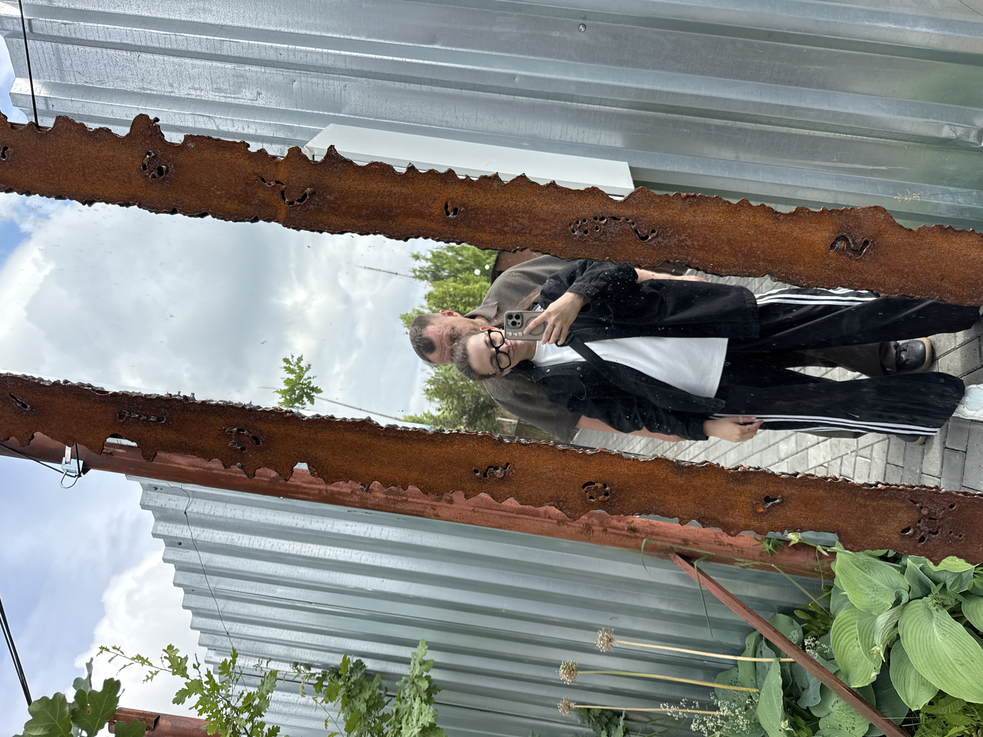 The image depicts a mirror reflection of two individuals standing in front of a corrugated metal fence and some greenery. In the foreground of the reflection, a young woman with glasses is taking a selfie with her phone. She is wearing a black jacket over a white t-shirt and black pants with white stripes. Behind her, a man with short, dark hair is visible. The background shows a cloudy sky, trees, and the texture of the metal fence. The mirror itself appears to be old and has a rusty, jagged frame. The setting is outdoors, possibly in an urban or industrial area, given the fence. The specific location in Izhevsk, Russia, is not discernible from the visual information provided in the image.