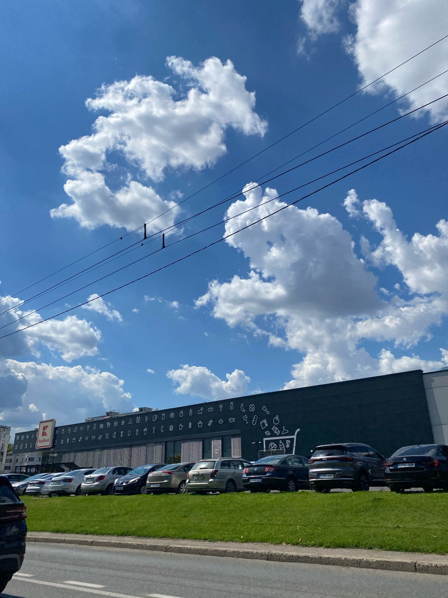 The image shows a view of a Kaufland supermarket in Chișinău, Moldova. The building has a dark facade decorated with white icons of various products. A parking lot filled with cars is in front of the supermarket, and a grassy median separates the parking lot from a road. The sky is blue with fluffy white clouds. Power lines are visible in the sky.