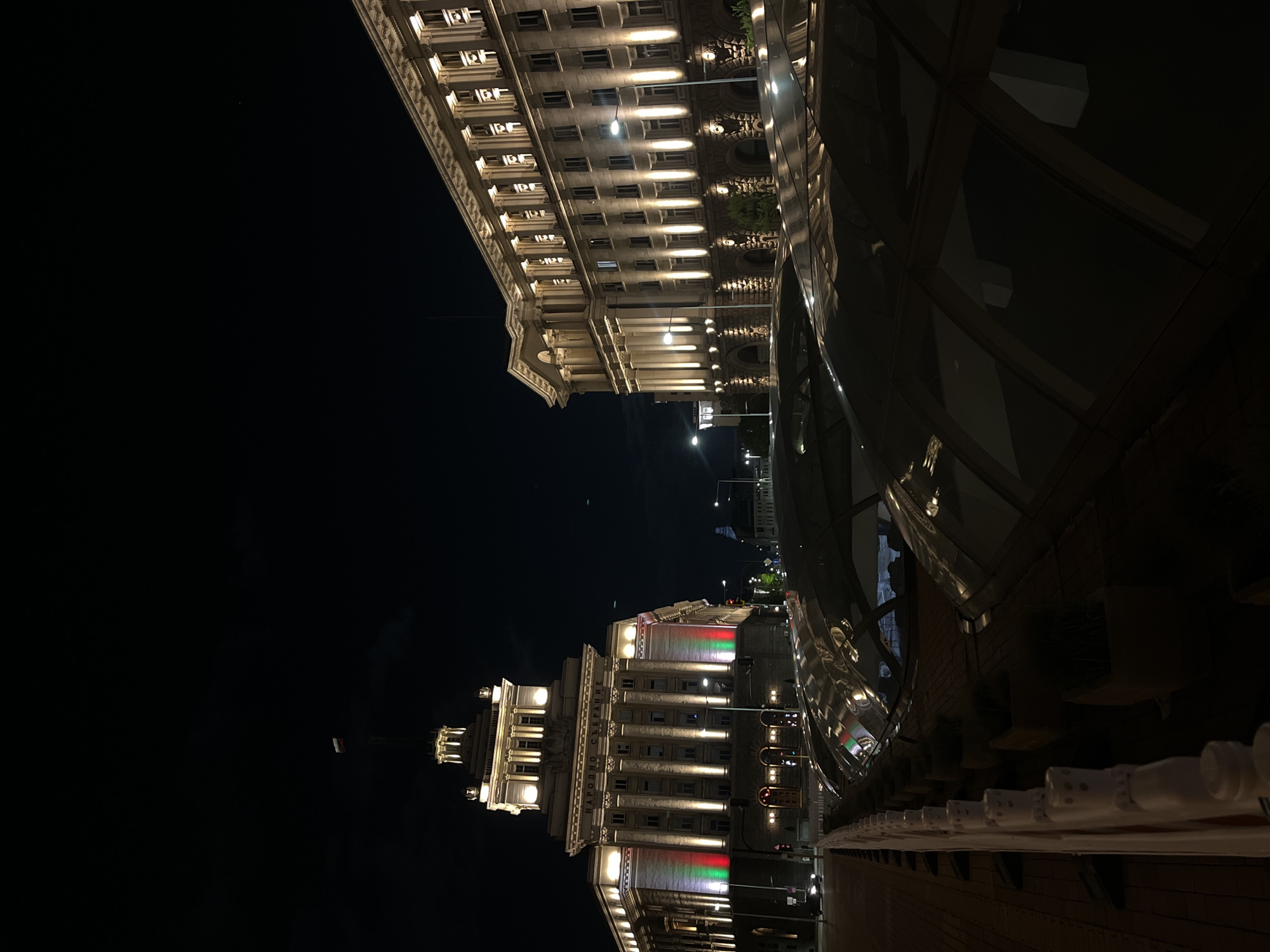 The image captures a striking nighttime cityscape in Sofia, Bulgaria, presenting grand illuminated architecture under a clear, dark sky. The scene is dominated by two impressive buildings, brilliantly lit to highlight their classical design.
On the left, the facade of a large, multi-story building is prominently featured, identified by the text "НАРОДНО СЪБРАНИЕ" (Narodno Sabranie), which translates to National Assembly or Parliament. This building boasts numerous pillars and windows, all accentuated by warm, upward-projecting lights. Notably, sections of its facade are illuminated with red, white, and green lights, subtly resembling the colors of the Bulgarian national flag.
Adjacent to this, further to the right and slightly set back, another large, ornate building extends, also brightly lit and characterized by arched windows and detailed architecture. In the immediate foreground on the right, a modern, curved glass structure with a highly reflective surface stands out. It mirrors the bright lights of the surrounding buildings and streetlights, creating dynamic patterns of light across its dark, sleek surface.
Streetlights are visible throughout the composition, adding further illumination to the urban environment. The sky above is a deep, unblemished black, indicating a clear night. A faint, small light source can be seen in the distant sky. The overall atmosphere conveys a sense of historical grandeur and vibrant urban life after dark in Sofia.