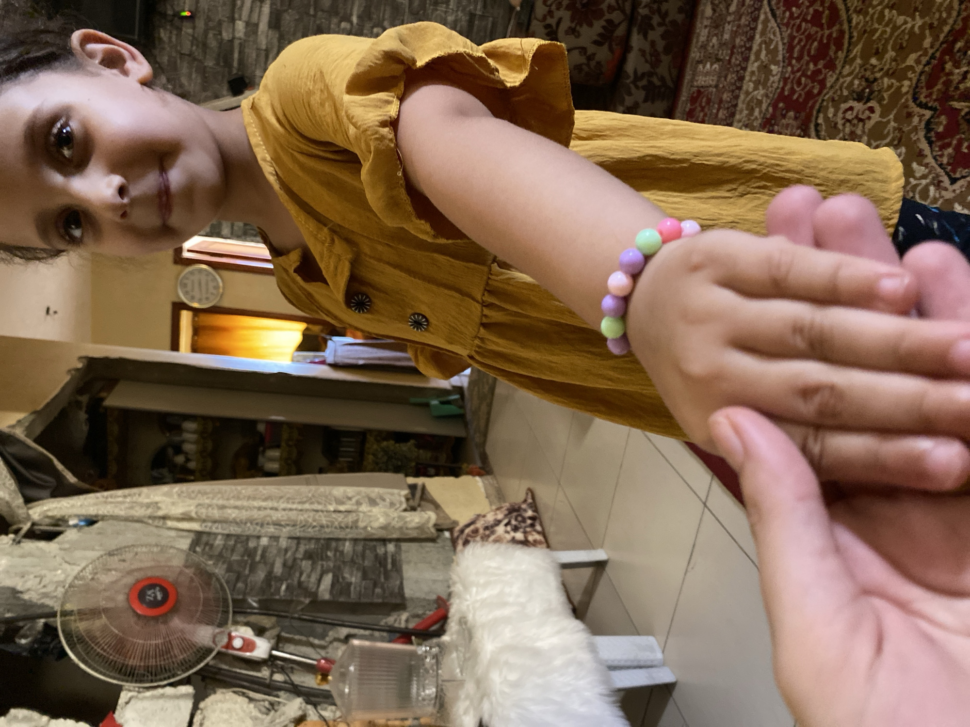 The media file is a close-up, slightly tilted shot of a young girl interacting with another person's hand. The girl, with dark hair and expressive eyes, is wearing a mustard-yellow dress with ruffled sleeves. She has a colorful beaded bracelet on her left wrist, featuring pastel-colored beads in shades of green, purple, pink, and orange. Her right hand is held out, and the other person's hand is gently touching it.
The background is somewhat blurred but reveals an indoor setting. To the left, a portion of a room with patterned wallpaper is visible, along with what appears to be a round clock on the wall. In the lower part of the image, there is a large, clear oscillating fan. The overall impression is one of a casual, intimate moment between two individuals, likely a parent and child, given the context of the location. The lighting suggests it might be daytime, but the indoor setting makes it difficult to determine definitively. The emotional tone conveyed is tender and playful. The location is specified as Khan Yunis, Palestinian Territories.