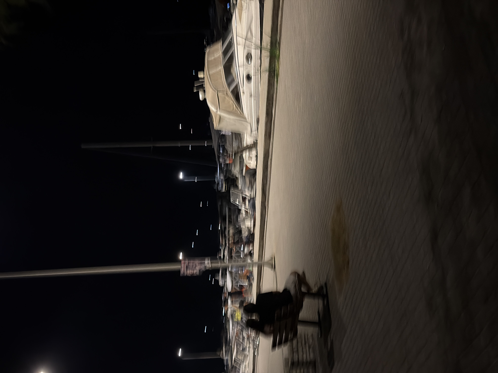 At night, two individuals are seated on a park bench, their backs to the camera, in the Municipal Unit of Alyzia, Greece. They are facing a body of water where numerous boats are docked. The scene is dimly lit by streetlights, casting long shadows. The boats appear to be a mix of recreational and possibly commercial vessels, with some covered and others exposed. The ground is paved with interlocking bricks, and there is a light post with a sign positioned behind the bench. The overall atmosphere is tranquil and serene, characteristic of a quiet evening at a marina.