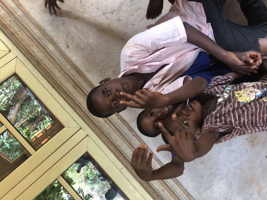 Three dark-skinned children pose on the floor near a window. The child at the top wears a pink shirt and dark pants. The second child has on a blue shirt. The third child is in a striped shirt and holds a lollipop in their mouth. All three are making hand gestures, showing two fingers in front of their eyes. Part of a wooden-framed window is visible on the left, reflecting trees and foliage. A partial hand and foot are visible in the top of the frame.