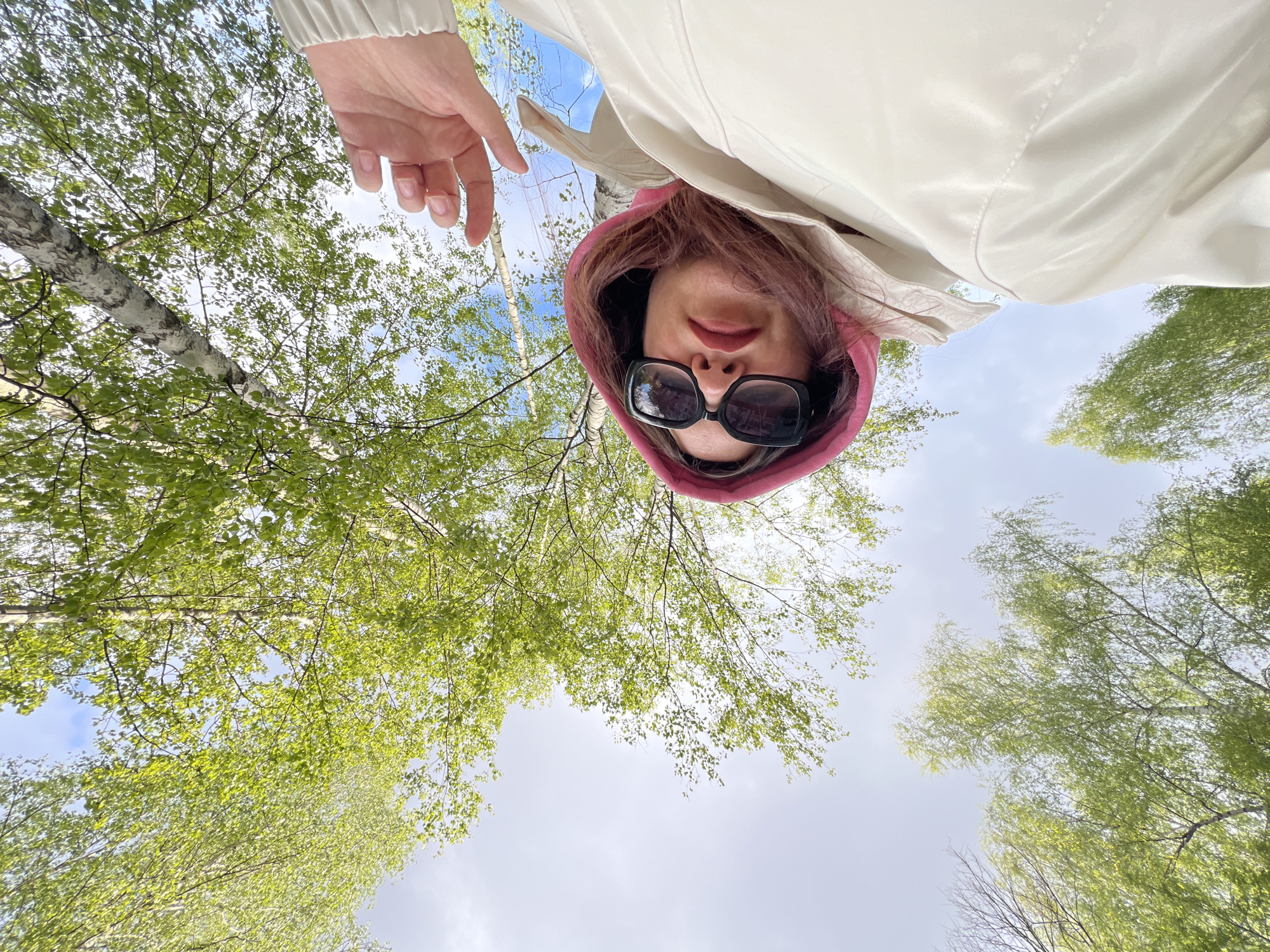 A person wearing a white jacket with a pink hood and sunglasses is looking down at the camera. A hand is visible near the top left of the frame. The background consists of trees with green leaves and a sky with light clouds. The picture appears to be taken from a low angle, looking up at the person. There is no clear evidence indicating the location to be Noviy, Russia.