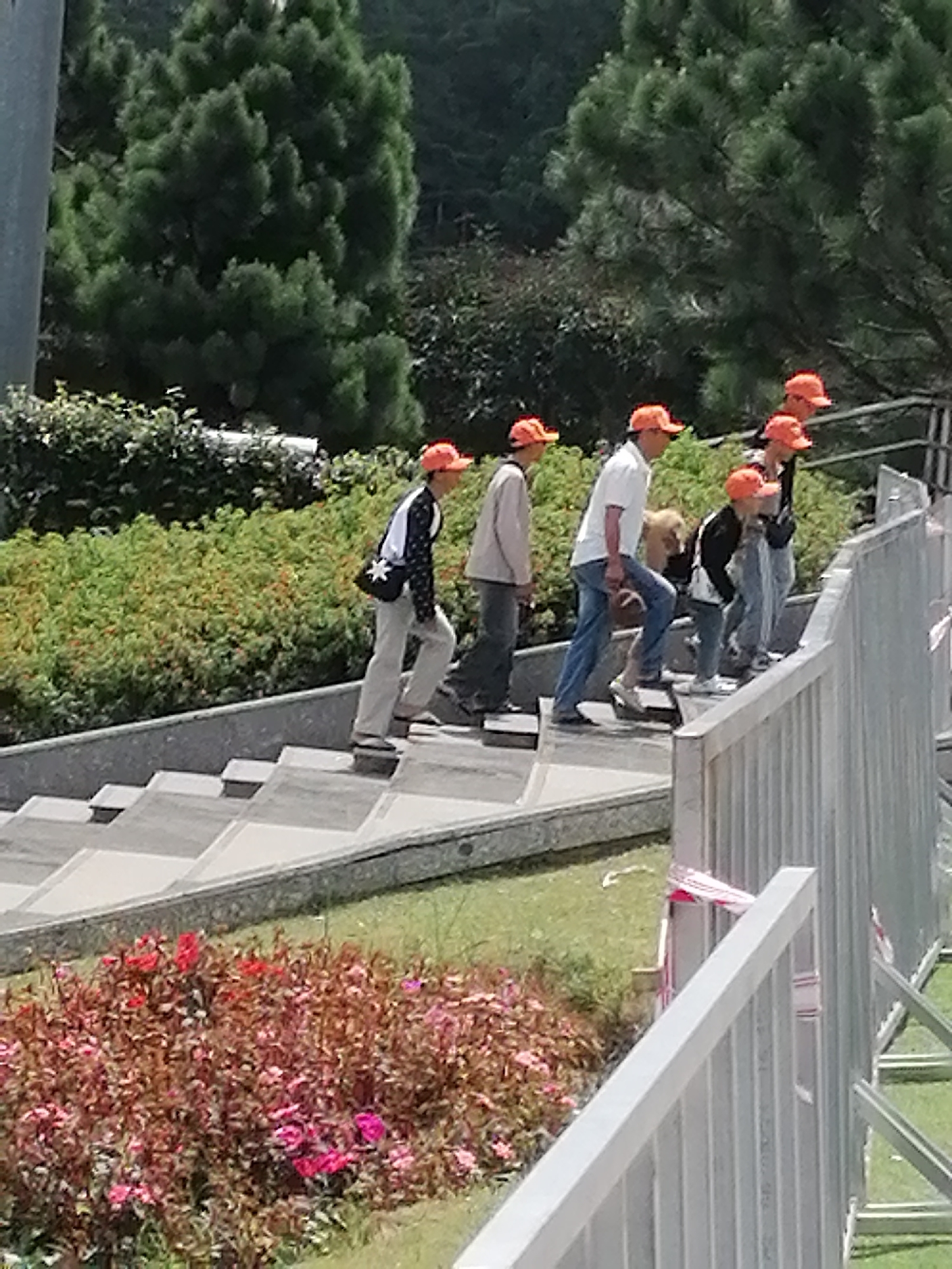 A group of people, mostly children wearing orange caps, ascend a wide, outdoor concrete staircase. The individuals are dressed casually in shirts, pants, and a mix of jackets and vests. Some carry bags. The staircase is bordered by lush green foliage and a grey metal railing on the right. In the background, large evergreen trees are visible. The scene appears to be a public space, possibly a park or a tourist attraction, in the city of Da Lat, Vietnam.