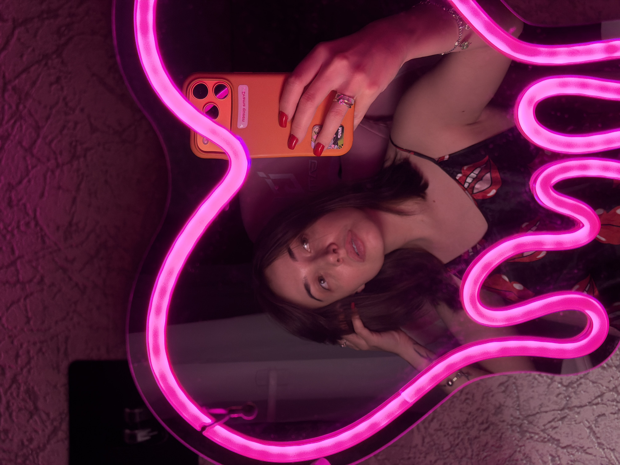 A young woman takes a mirror selfie in a dimly lit room. She is reflected in a heart-shaped mirror with pink neon lights. She is wearing a black slip dress with a red lips pattern and her nails are painted red. She has a ring on her left hand and a bracelet on her right wrist. She is holding her phone, which has an orange case, in her right hand. The room appears to have textured walls.