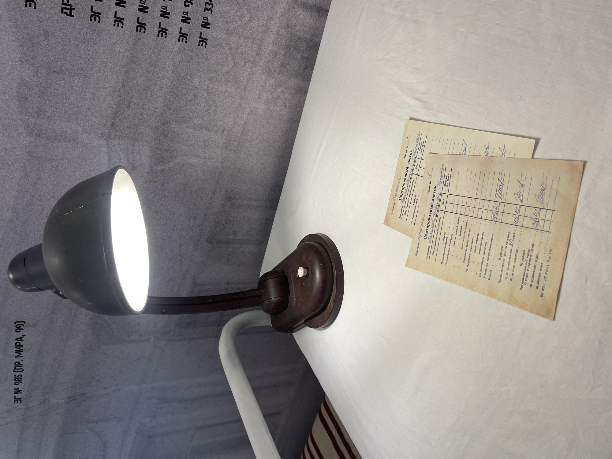 A desk lamp illuminates two forms in Russian on a white surface. The lamp is dark gray with a brown mounting base, and the bulb is lit. The papers are titled "Sortirovochny Listok" (Sorting Sheet). The background includes gray fabric with printed text "ЭГ N 985". The location of the city Krasnoyarsk, Russia is not discernable in the image.