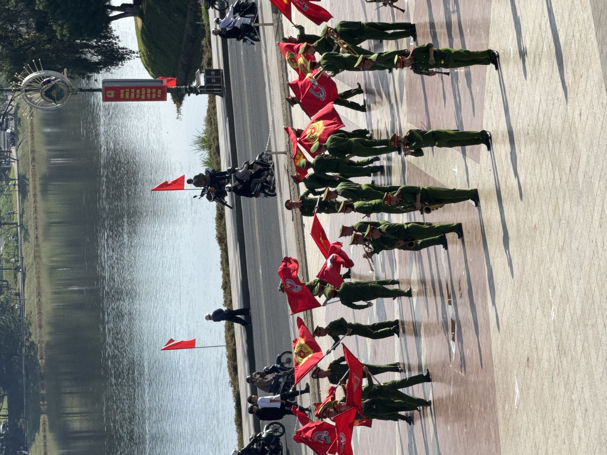 The image depicts a sunny outdoor scene with a body of water in the background, likely Xuan Huong Lake in Da Lat, Vietnam, given the presence of a lamppost with a municipal emblem and signage. In the foreground, a group of men in matching green uniforms with red caps are positioned on a paved square, some holding red flags with yellow emblems and others holding musical instruments. Several individuals are playing brass and woodwind instruments, suggesting a marching band or ceremonial band. A few people on motorcycles are visible on a road behind the square, and a lone figure in a dark outfit is walking away from the viewer near the water's edge. The overall atmosphere appears to be a public event or ceremony.