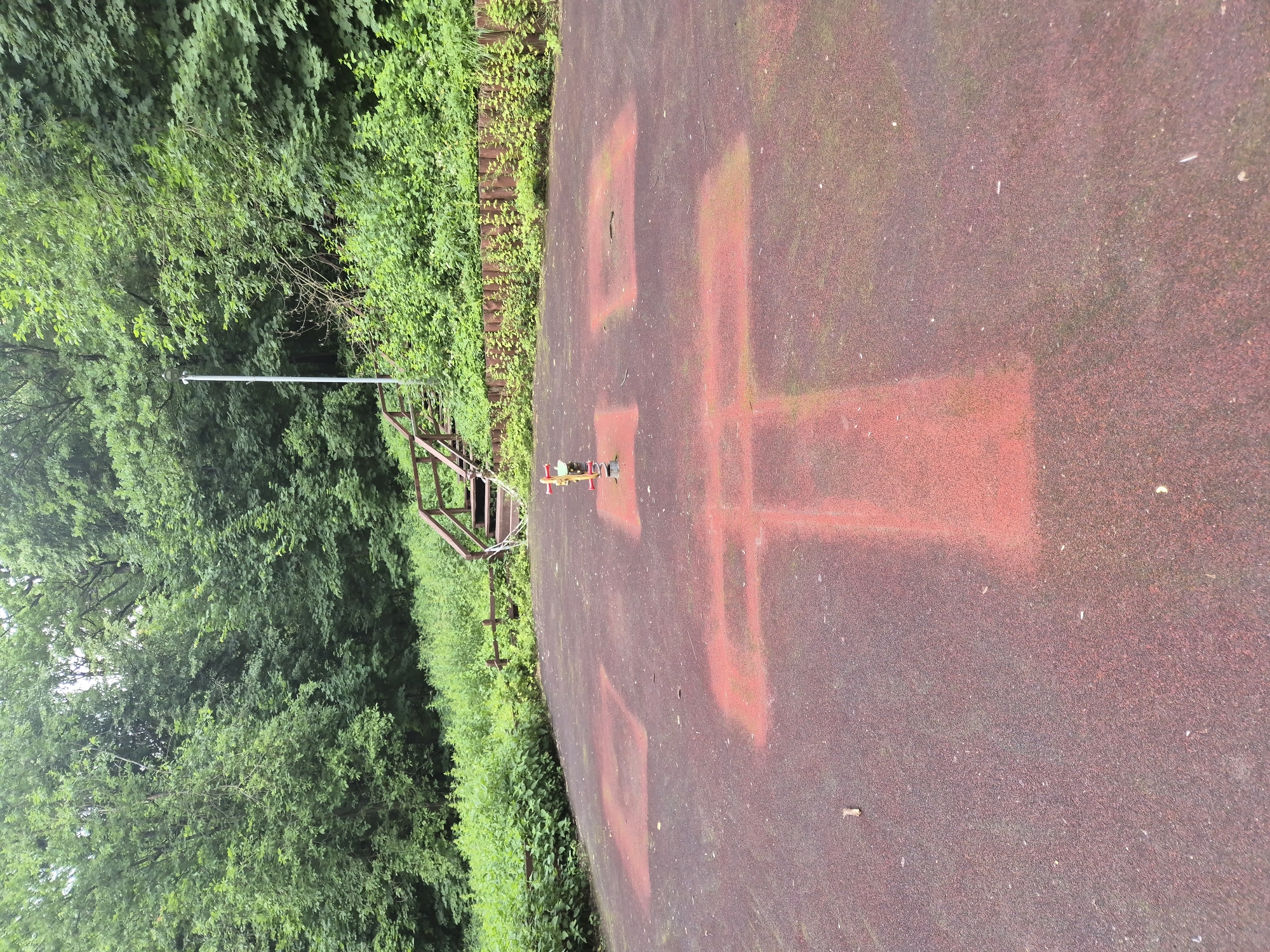 A playground is located in a forested area in Moscow, Russia. The playground surface is a dark red rubberized material with faded red markings. In the center of the playground, there is a single piece of red and yellow playground equipment. In the background, a wooden staircase leads up a grassy hill covered in dense green foliage and trees.