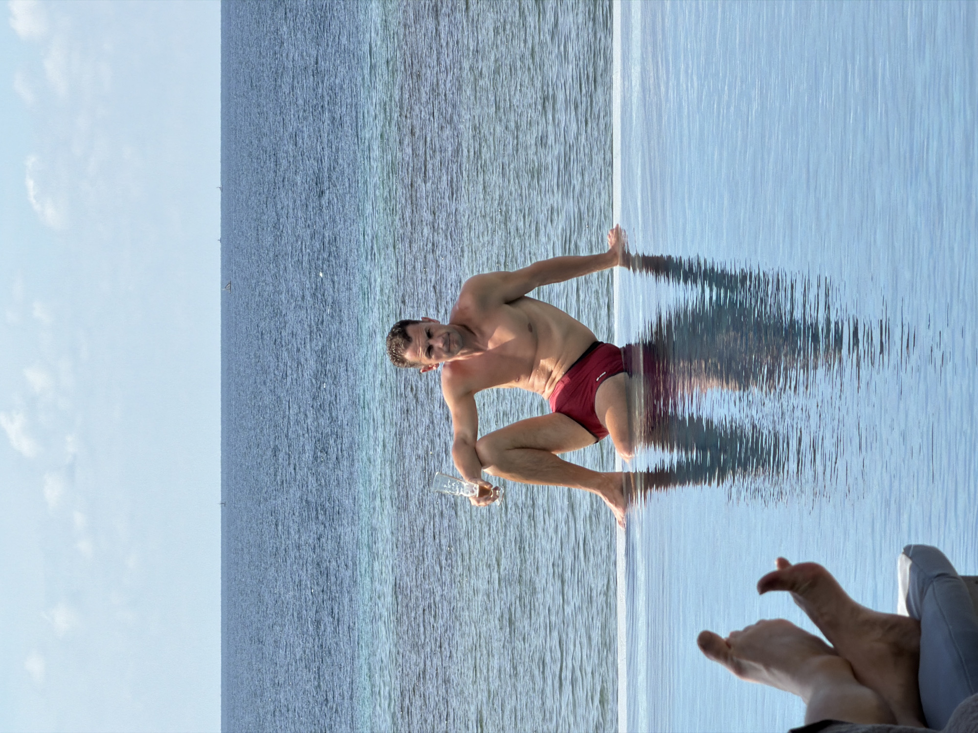 A man in red swimming trunks is crouched at the edge of an infinity pool, holding a glass in his left hand. His right hand rests on the edge of the pool. The pool water blends seamlessly with the ocean beyond, which stretches to the horizon under a bright sky. In the foreground, feet are visible, presumably belonging to someone relaxing. The location is Unknown, Indonesia.