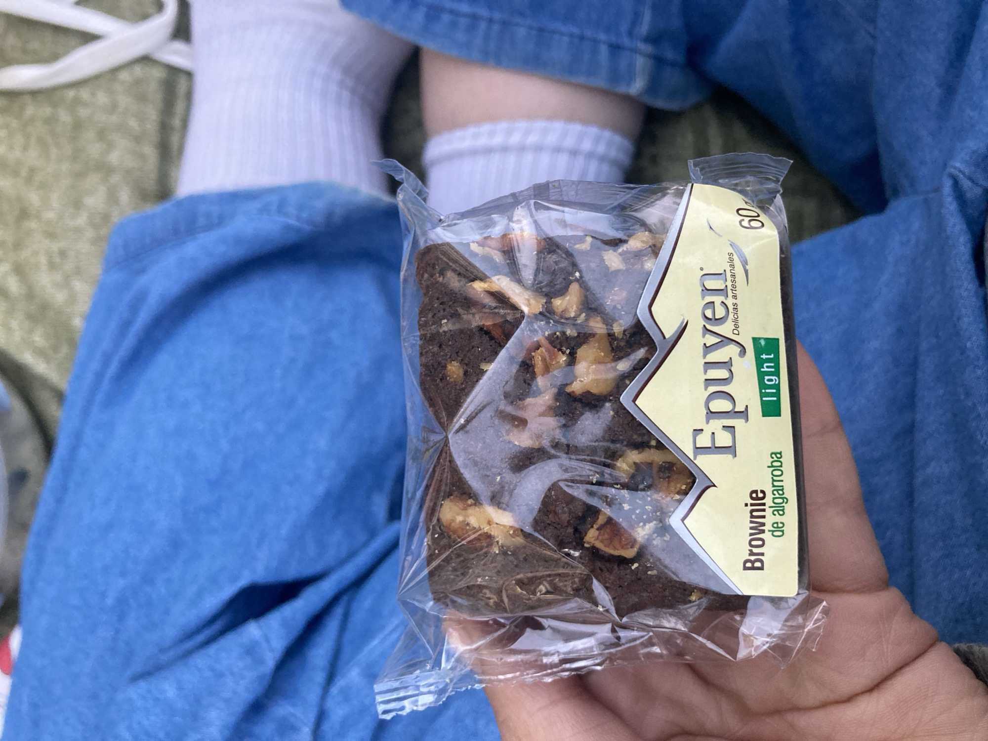 A person, likely in Buenos Aires, Argentina, is captured holding a single-serving packaged brownie. The focus is on the brownie and the hand holding it, with the person's legs and feet visible in the background. They are wearing light blue denim jeans and white ribbed socks, suggesting a casual and relaxed environment.
The brownie itself is a dark brown, rectangular treat, generously topped with visible pieces of nuts, possibly walnuts or pecans. It is encased in clear plastic packaging. The packaging features a prominent yellow label with green accents. Visible text on the label includes the brand name "Epuyen" and the product description "Brownie de algarroba" (carob brownie). Below this, the word "light" is displayed in a small green box. Other phrases on the label are "Delicias artesanales" (artisan delicacies) and the number "60," which could indicate the weight in grams or caloric content.
The setting appears to be outdoors or in a very casual indoor space, with a textured, greenish surface visible beneath the person's legs, possibly grass or a blanket. The lighting is bright and consistent with daytime. The overall scene suggests a moment of rest or a snack break.