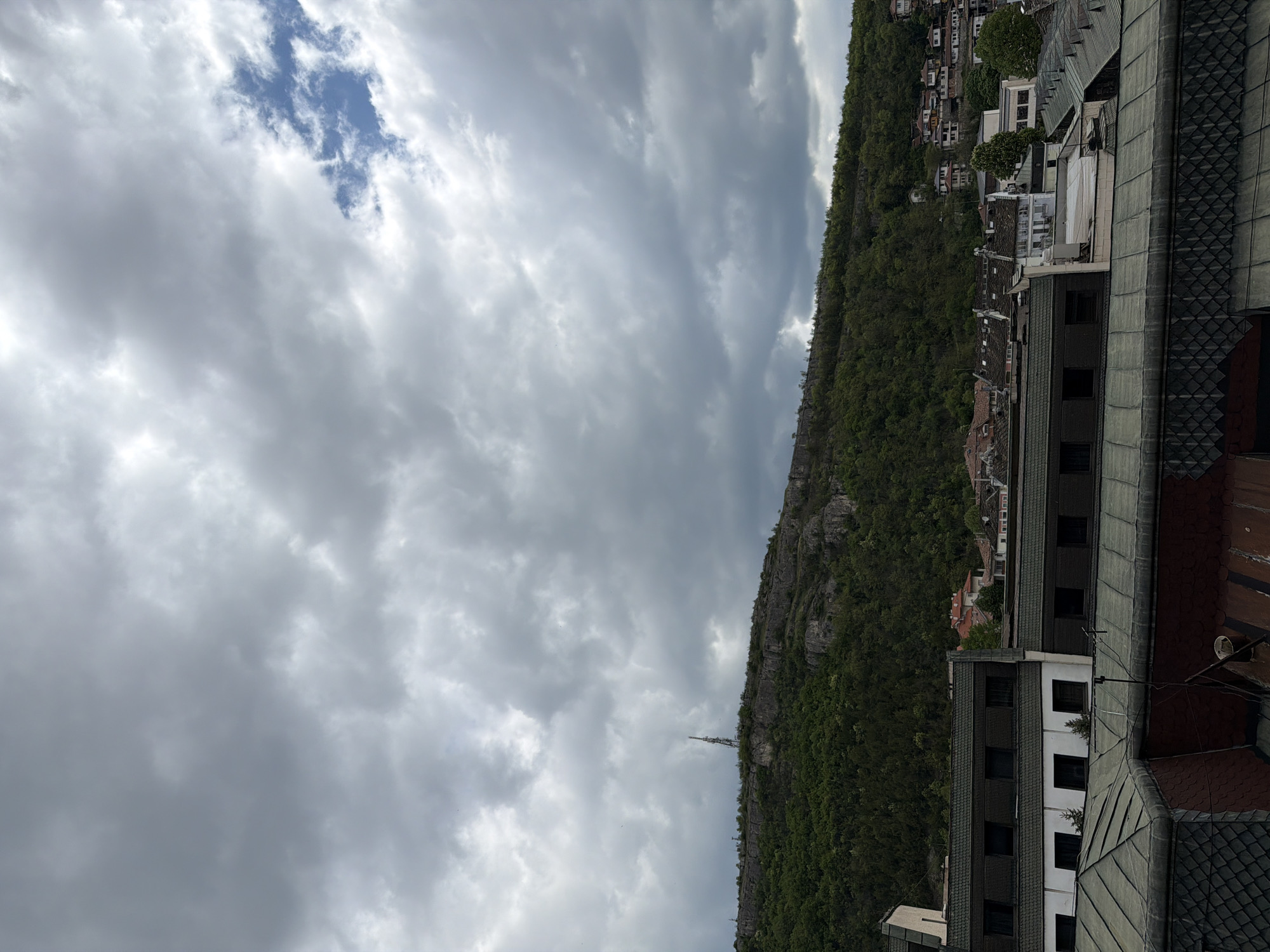 The image shows a view in Lovech, Bulgaria, looking across a cliff covered in trees and houses. The sky is overcast with clouds. A radio tower is visible on the cliff edge. The foreground includes buildings with tile roofs and windows. The buildings are of various heights and colors.