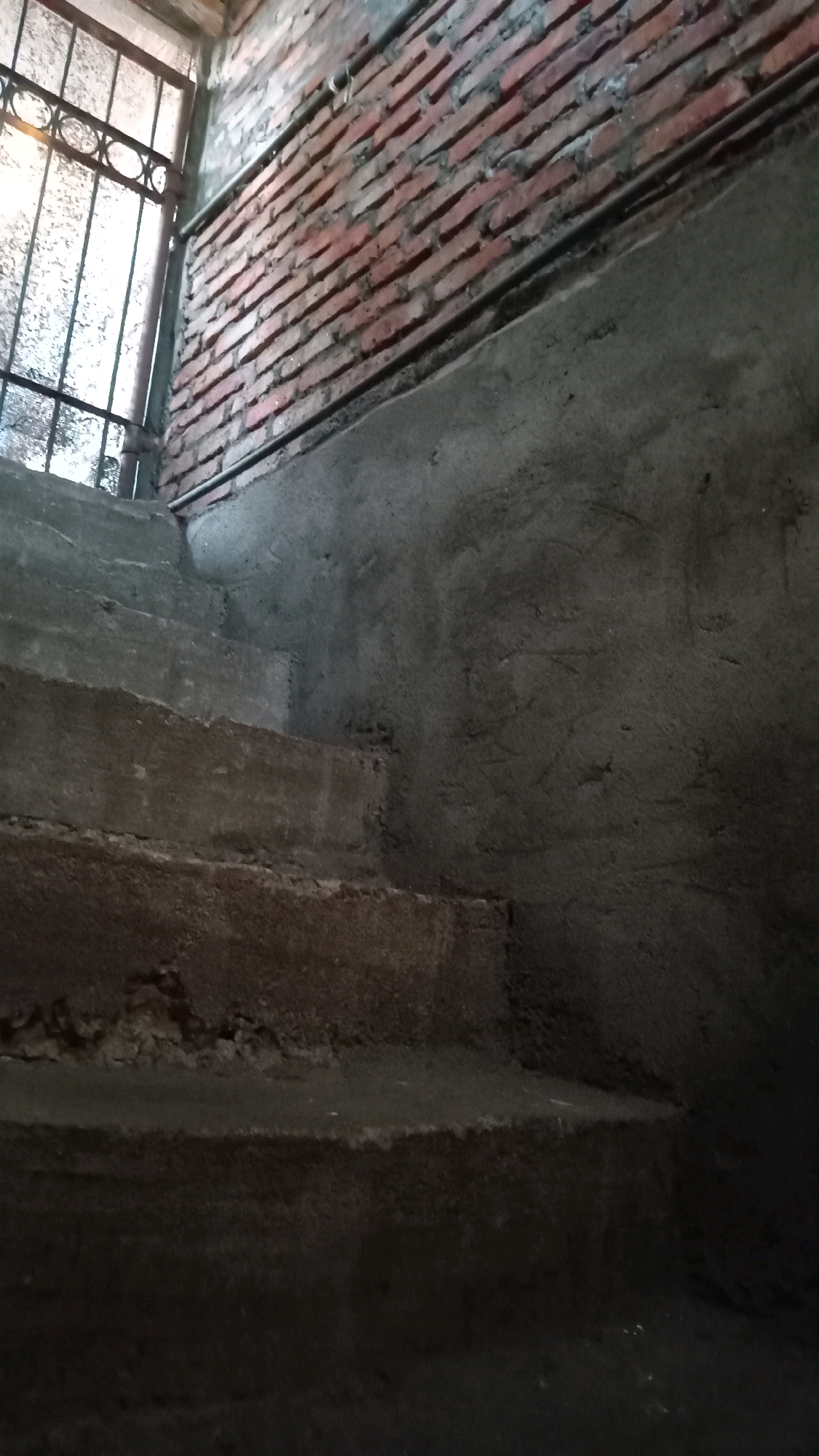 The image depicts a dimly lit, unfinished concrete staircase leading upwards. To the left, a metal grate covers an opening, with faint light filtering through. The wall behind the grate is constructed of red bricks, with visible mortar. Adjacent to the staircase, the wall is covered in rough, gray concrete. The overall scene suggests an interior space, possibly a stairwell or an outdoor path under construction or in a state of disrepair. The location is identified as Randuagung, Indonesia. There are no people or animals visible in the image.