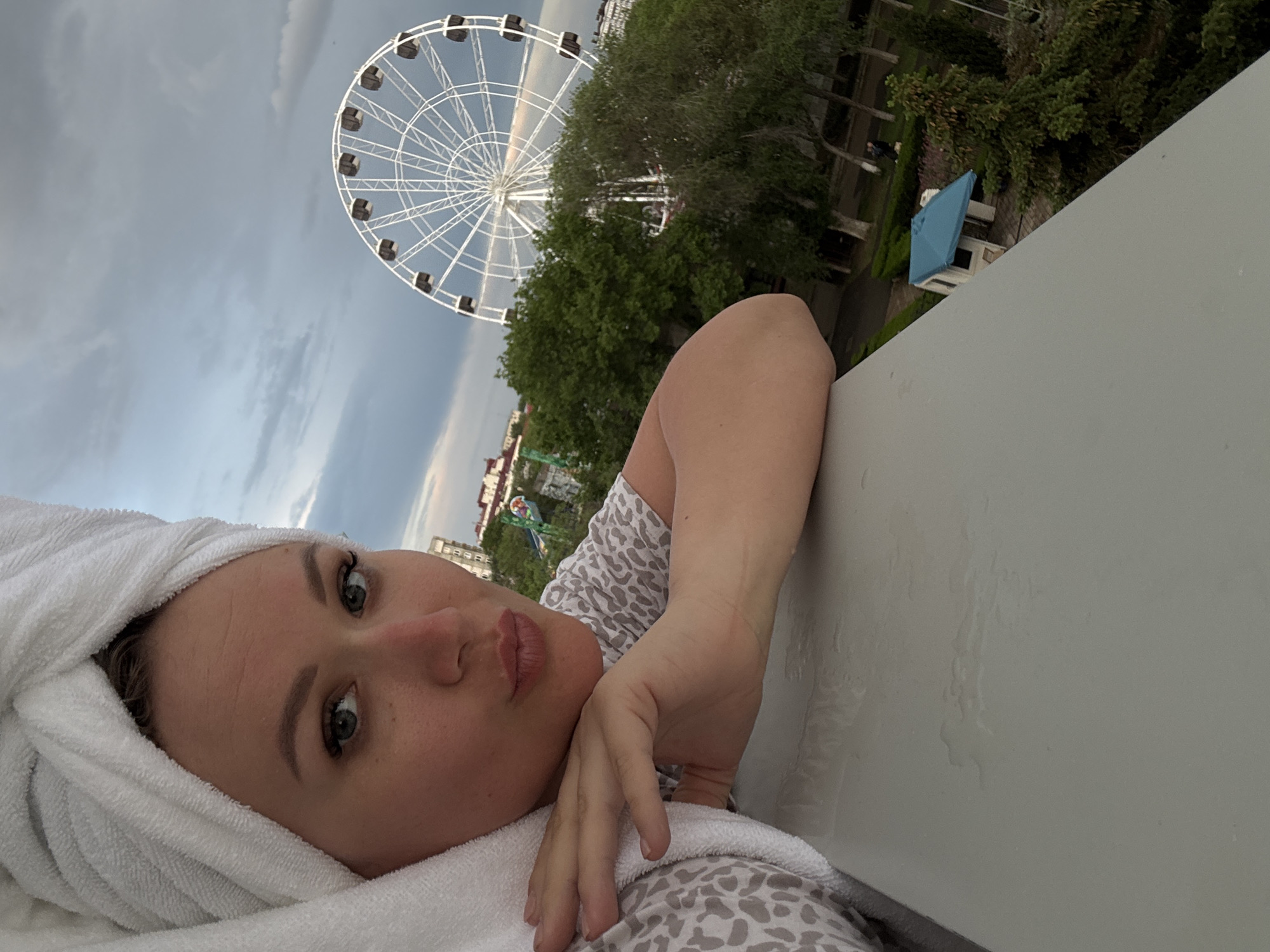The photograph shows a woman with a towel wrapped around her head, resting her head on her arm which is placed on a gray, flat surface. The woman is fair-skinned with light-colored eyes. The background includes a Ferris wheel and trees. A small blue and white structure is also visible in the background. The photograph appears to be taken at an angle, which rotates the traditional vertical view. The location is in the city of Yevpatoriya, Ukraine.