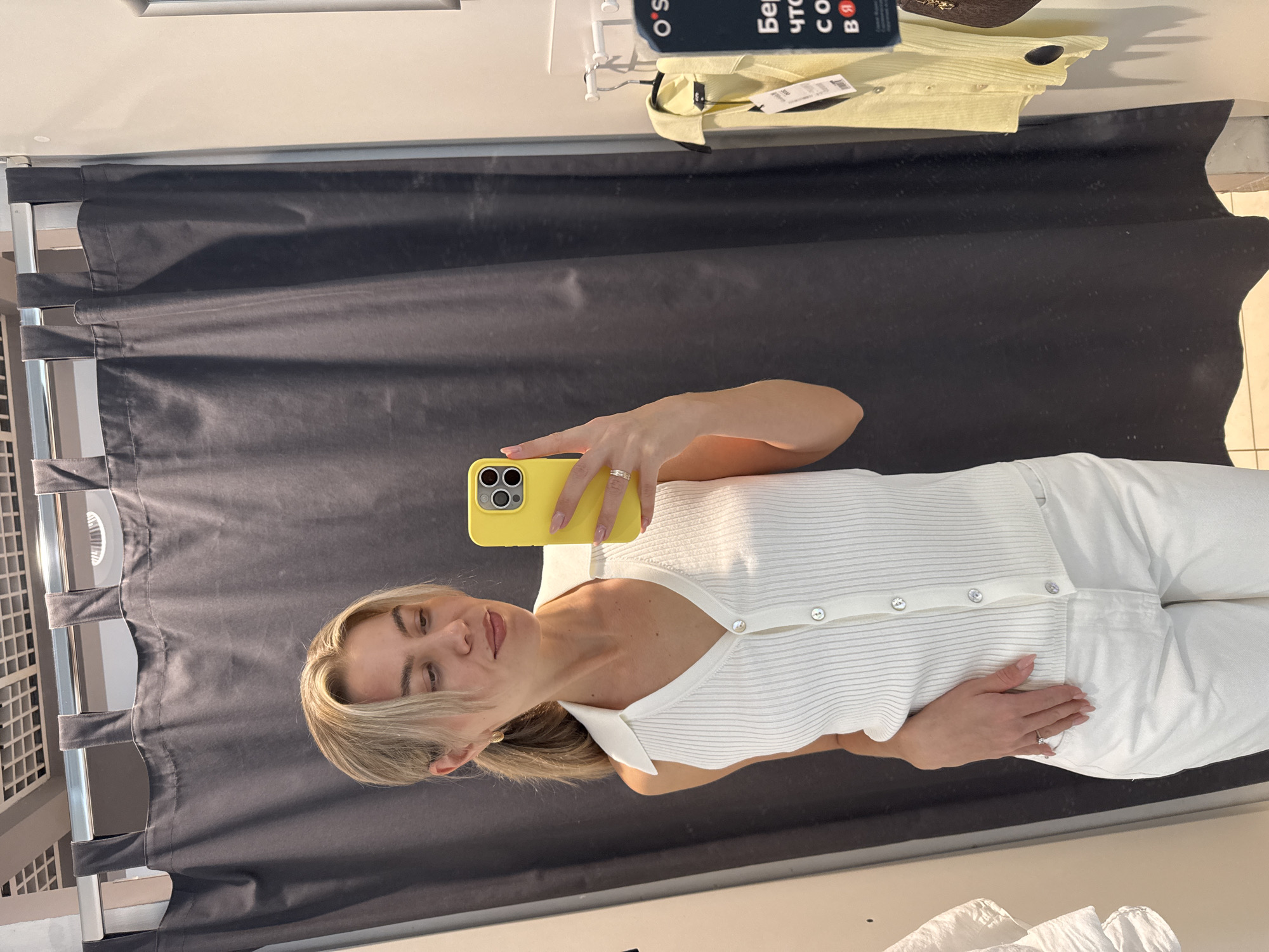 A woman stands in a dressing room, holding a yellow smartphone in her right hand as she takes a selfie in a full-length mirror. She is wearing a white ribbed top with buttons and white pants. The dressing room features a dark gray curtain backdrop and the metal structure for the curtain. In the upper part of the frame, a yellow garment hangs on a hanger, along with a tag written in Cyrillic script. The specific location, city, and country are not identifiable in the provided image.