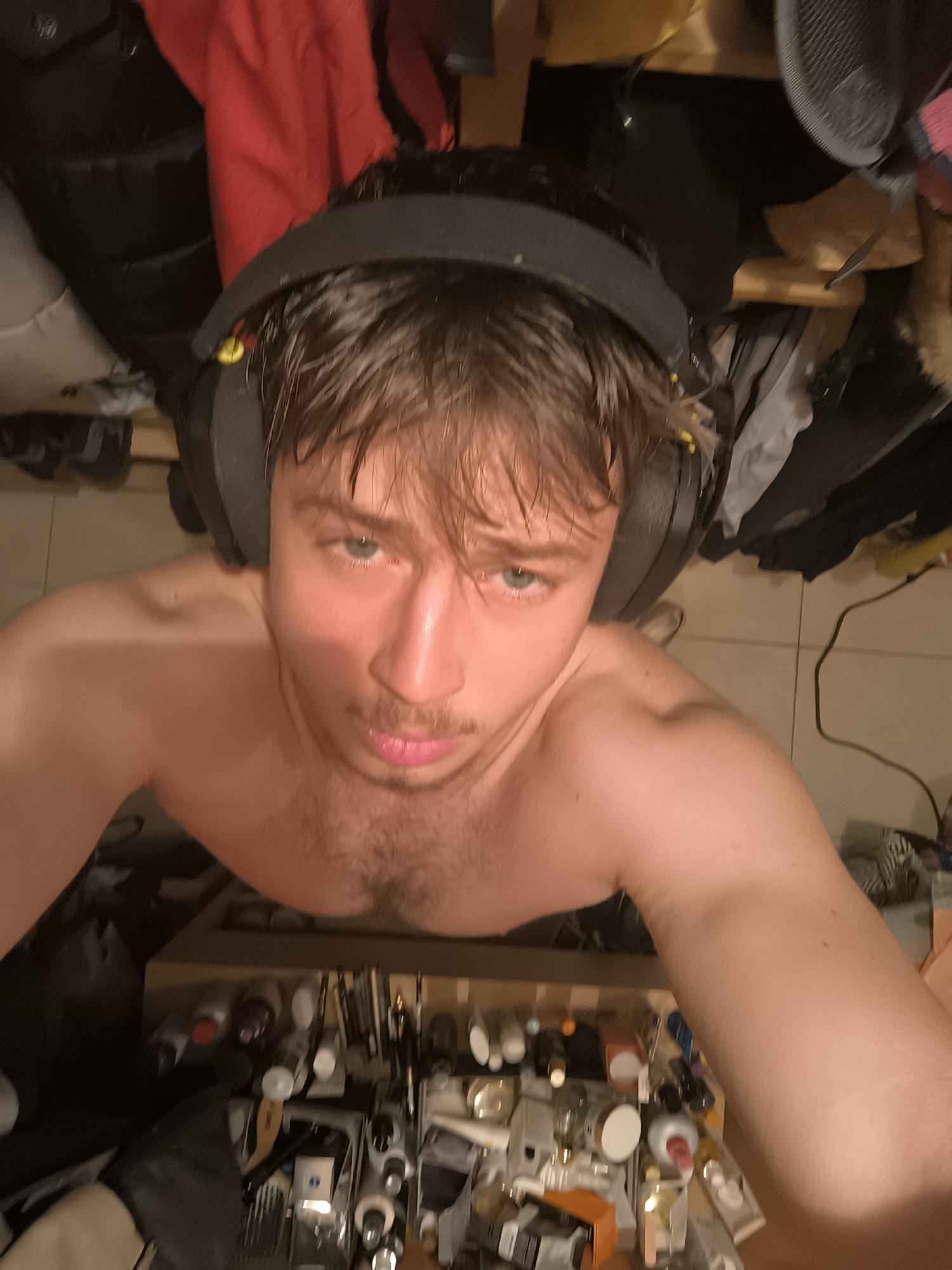 A low-angle, close-up selfie shows a shirtless young man wearing black headphones. He has short, brown hair and a light mustache. His chest is visible and shows some hair. Behind him, and slightly out of focus, is a cluttered area containing items like bottles, jars, and makeup brushes, possibly a vanity or storage area. Further behind, there are clothes hanging. The location, Sofia, Bulgaria, is not discernible from the visual information provided in the image.