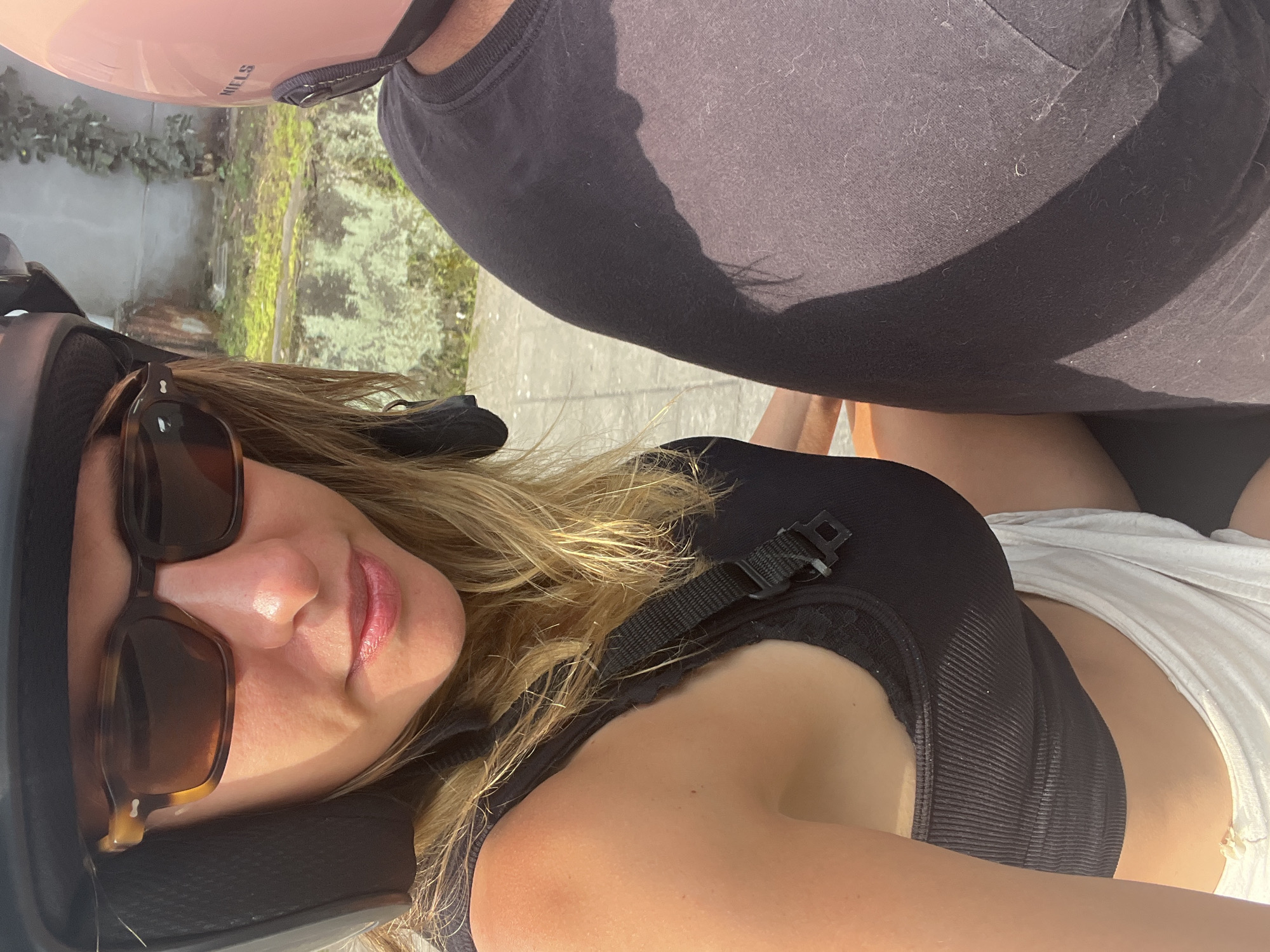 A close-up, waist-up, outdoor, selfie-style photograph shows two individuals on a motorcycle, viewed from the perspective of the passenger. The passenger, on the left, is a Caucasian woman with blonde hair, wearing a black helmet, tortoiseshell sunglasses, a black ribbed tank top, and light-colored shorts. Her left arm is visible, and she is looking towards the camera. The driver, on the right, is wearing a black t-shirt and a pink helmet. The background consists of a weathered concrete wall with green foliage climbing it, and a patch of ground covered in moss and debris. The lighting suggests it is daytime, with strong sunlight casting shadows. The image quality is consistent with a smartphone camera.