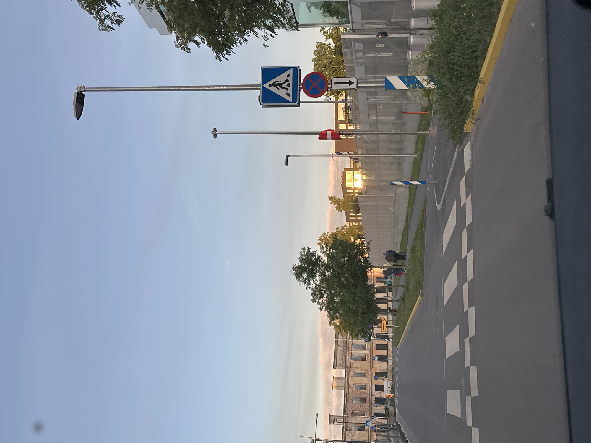 The image shows a street in Tallinn, Estonia, with an old building in the background and modern structures. The foreground features a pedestrian crossing, traffic signs, and streetlights. The sky is clear with a few clouds. In the middle ground, there are trees, a fence, and some people walking. The lighting suggests it's either early morning or late afternoon.