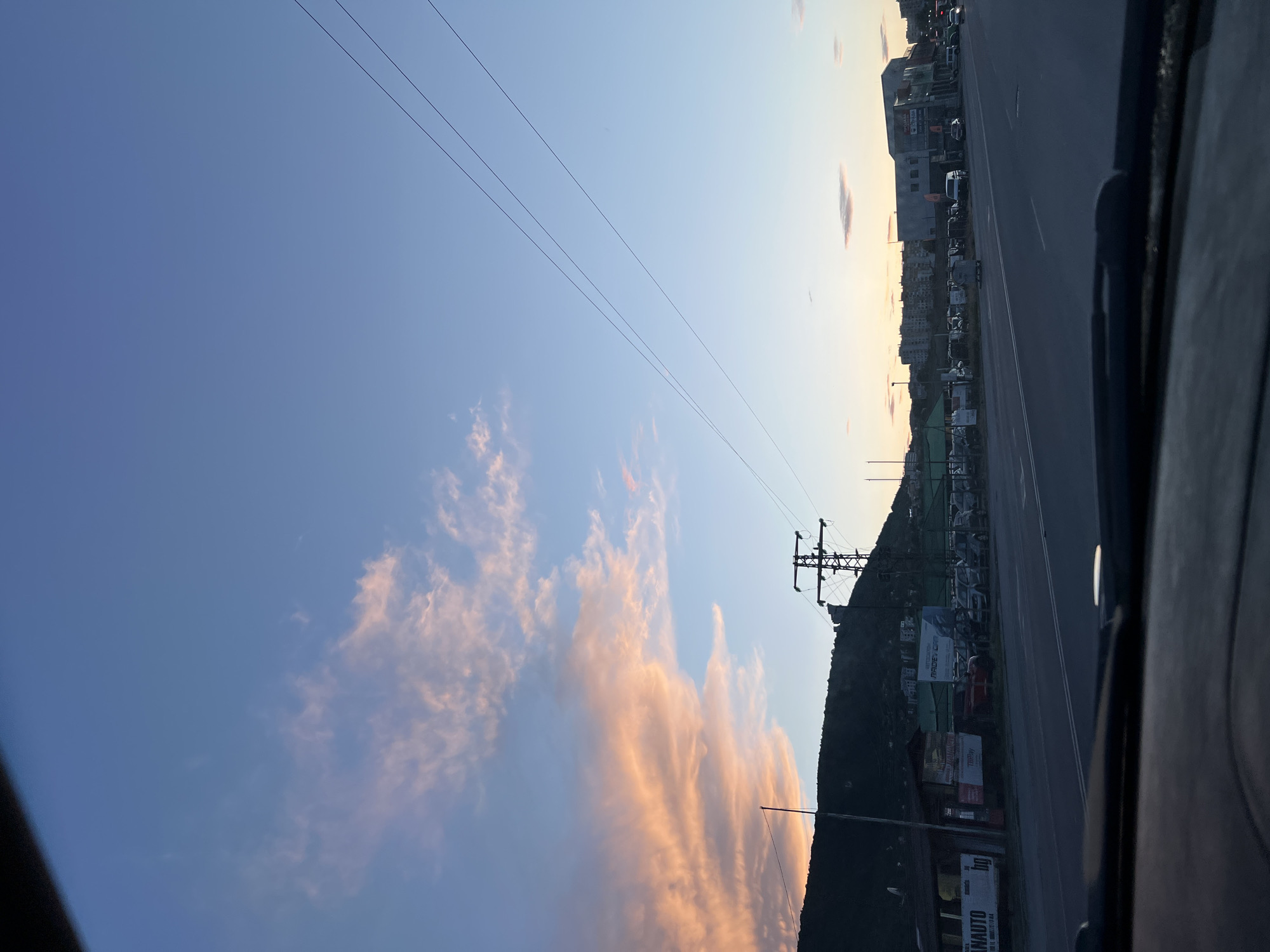 The image displays a roadside view at either sunrise or sunset, characterized by a dramatic sky and an urban fringe landscape. The sky, occupying the majority of the frame, transitions from a deep blue at the top to vibrant hues of light blue, yellow, orange, and pink towards the horizon, where elongated, wispy clouds are illuminated by the low sun. Several thin power lines stretch across the upper part of the sky.
Below the sky, a multi-lane paved road runs horizontally, leading into the distance. To the right of the road, a dark, undulating hill or mountain forms a prominent silhouette against the colorful sky. Along the roadside, various commercial buildings, businesses, and parked vehicles are visible. A large metal power pylon or transmission tower stands out on the right, silhouetted against the sky. The rightmost edge of the image shows a dark frame, suggesting the photo was taken from inside a vehicle.
Visible text on roadside signs includes "MAKBER CAR," "TRILPAY," and "AMAUTO." Another sign displays an "hp" logo and "SERVICIO TEC..." The time of day is clearly indicated as either dawn or dusk by the warm, soft lighting and the colors of the clouds. The weather appears clear with scattered clouds.