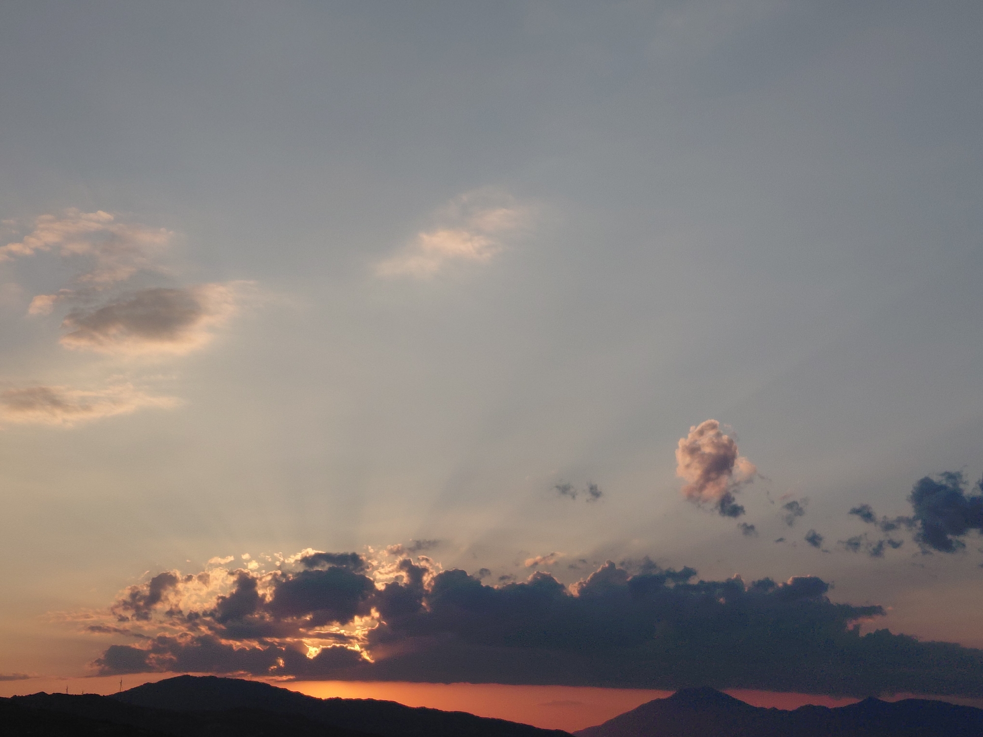 The image shows a sunset over a mountainous landscape. The sky is a gradient of gray and pink hues. Several clouds are present, some are dark and heavy, while others are lighter and wispy. Rays of sunlight are visible, breaking through the clouds and illuminating parts of the sky. The lower portion of the image depicts a silhouetted mountain range, with hints of orange light peeking through the gaps. The scene appears to be taken from a distance, looking towards the horizon. The Municipal Unit of Aegio, Greece is characterized by its coastal mountains that meet the Gulf of Corinth, a landscape that aligns with the visual elements in the photograph. There are no people or specific structures visible that would indicate a precise urban location within Aegio itself. The image captures a natural, atmospheric phenomenon.