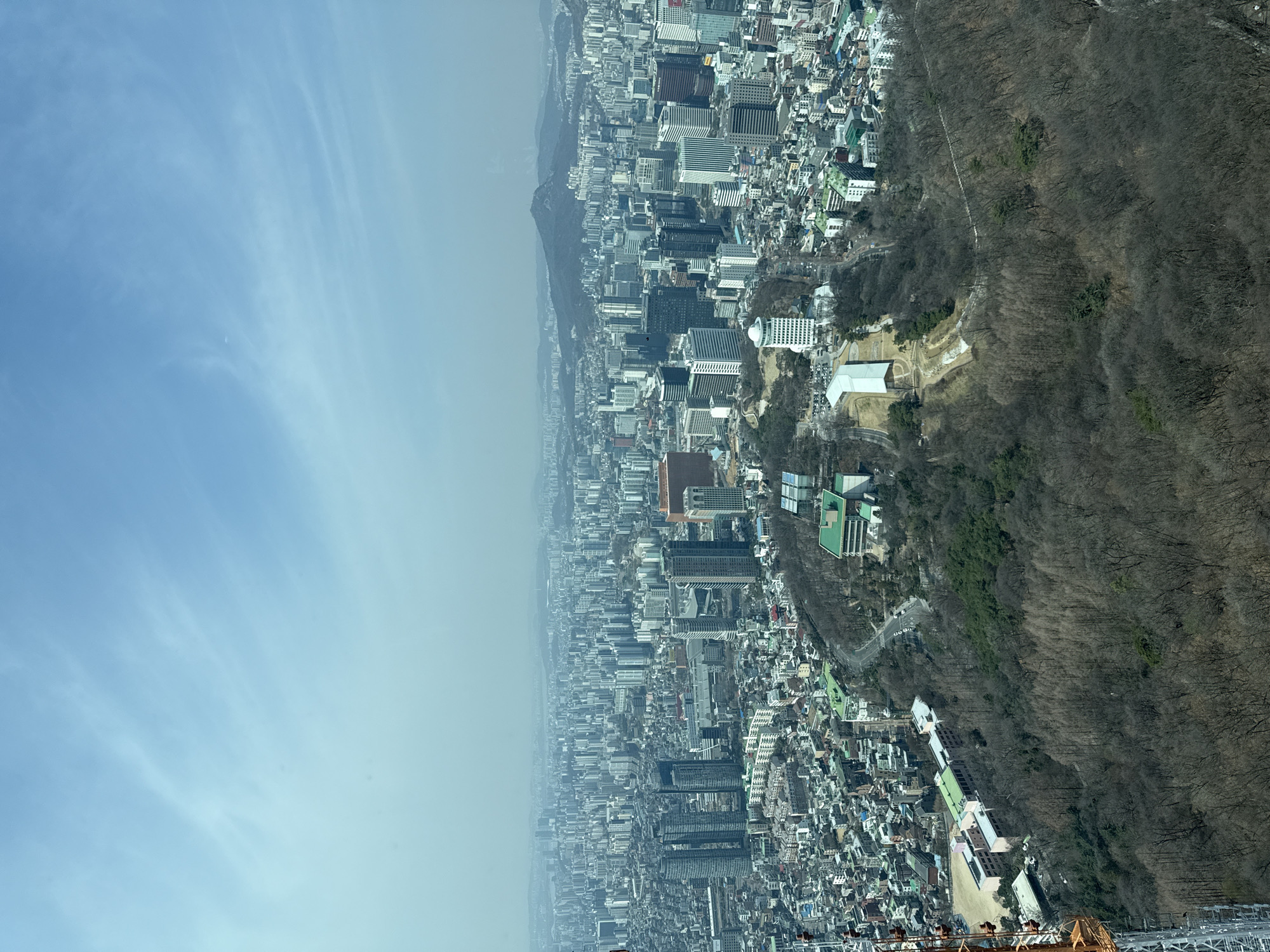 This high-altitude panoramic image depicts the dense urban landscape of Seoul, South Korea, viewed under a hazy blue sky with scattered wispy clouds. The perspective originates from an elevated structure on Namsan, likely the N Seoul Tower itself, looking generally northwest over Namsan Park and the surrounding city.
In the immediate foreground and extending into the mid-ground, the image is dominated by the largely deciduous, bare-treed slopes of Namsan Park, indicating late winter or early spring. A section of a large, lattice-structured communication tower, predominantly orange and white, is visible along the far left edge of the frame, extending upwards from the bottom.
Within the forested areas and on the park's slopes, several buildings are present:
* On the left, near the base of the visible tower, a complex of buildings with light pink facades and green roofs is visible adjacent to a cleared, dirt-colored area resembling a sports field.
* Towards the center, within the park, a distinctive white building with a large, curved, single-span roof is visible. Further up the slope, another prominent white building with a multi-tiered circular top is present. Other light-colored buildings with green roofs are scattered throughout the park.
* Winding roads are observed cutting through the forested sections, transitioning into the urban grid.
Beyond the park, the cityscape of Seoul sprawls extensively across the mid-ground and distant background. This area is characterized by an extremely high density of mid-rise and high-rise buildings, displaying a mix of residential and commercial architecture in various shades of grey, white, and darker tones. The urban fabric appears tightly packed, with roads and infrastructure interwoven between building clusters. In the far distance, the city merges into the horizon, rendered indistinct by atmospheric haze, with distant mountains faintly visible on the right. No individual people or specific actions are discernible due to the elevated vantage point and distance.