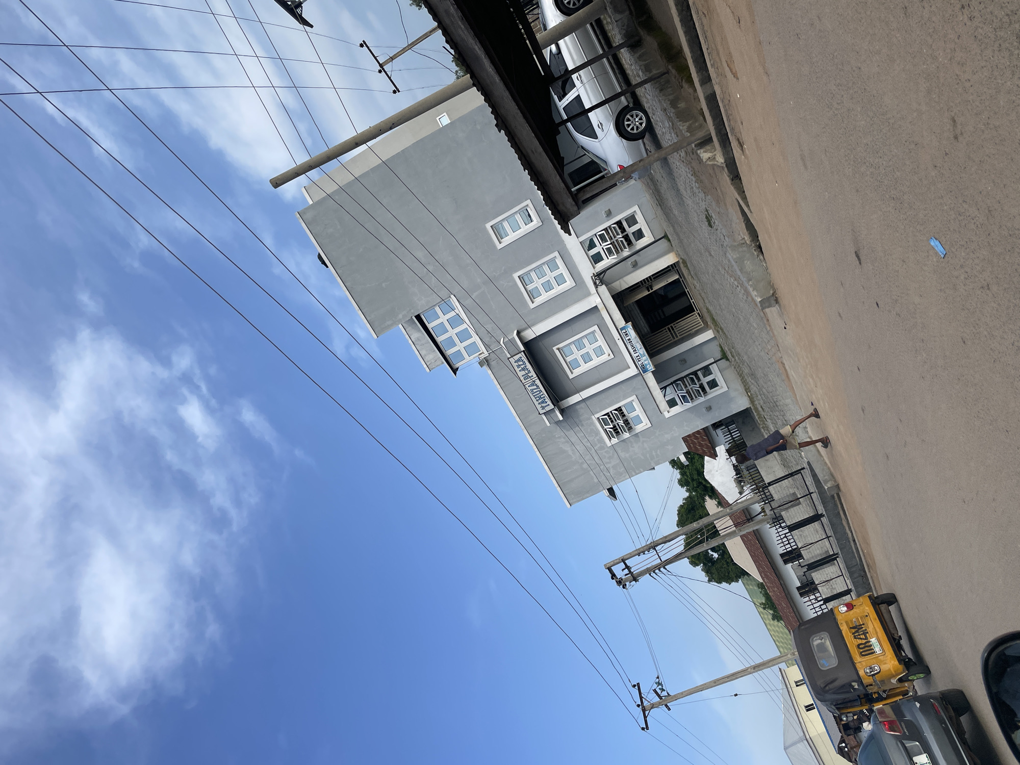 This image depicts a street scene in Jalingo, Nigeria. The primary subject is a grey, multi-story building with numerous windows, situated next to a paved road. A person is visible on the sidewalk in front of the building, walking away from the camera.
To the left of the building, a clear blue sky with some wispy clouds is visible, suggesting daytime. Electrical wires are strung across the scene, supported by utility poles. In the foreground, a yellow auto-rickshaw, also known as a Keke Napep, is parked on the side of the road, and a dark-colored car is partially visible behind it.
A sign on the building reads "TAHUZA PLAZA," and another sign, partially obscured, appears to be related to fashion. The overall setting appears to be a commercial or residential area with some overhead power lines. The lighting suggests it is either morning or late afternoon. The presence of the auto-rickshaw and the style of the building indicate a location in West Africa.