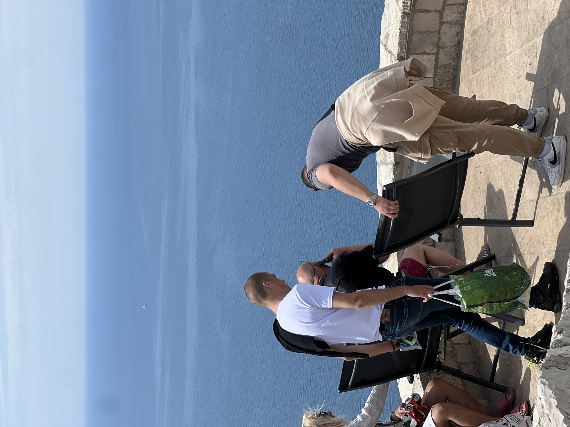 The image depicts a group of people on a stone patio overlooking a blue sea, likely taken in Sveti Stefan, Montenegro. Two people are seated on black chairs, one wearing a white shirt and jeans, and the other wearing a black shirt and red shorts. A green shopping bag is placed next to them. Another person is leaning over a chair, dressed in a gray shirt, tan pants, and a light-colored jacket. Their posture suggests they are looking down at something below. The sky is light blue and clear.