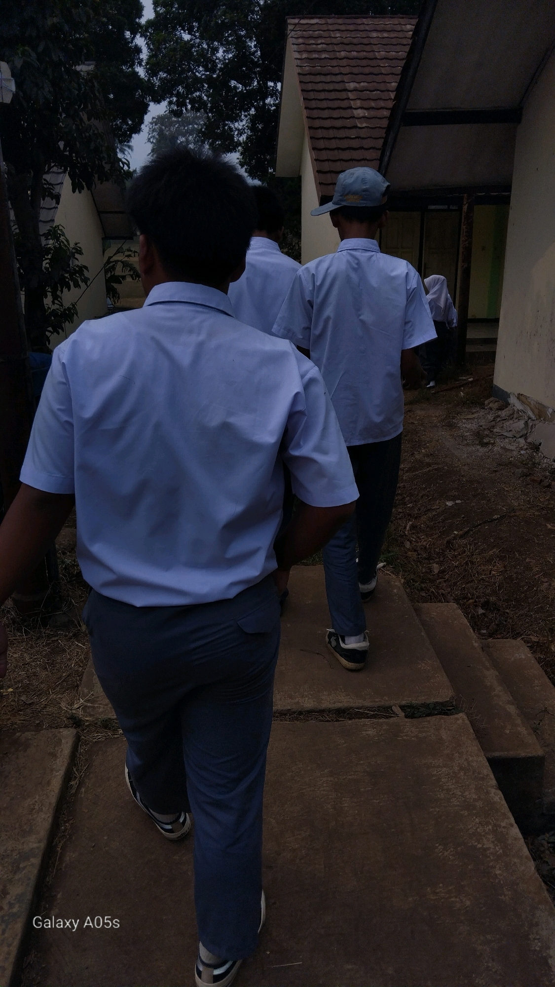 In Sumedang, Indonesia, three young individuals, presumably students, walk away from the camera down a concrete path. They are all wearing similar light blue short-sleeved shirts and dark grey trousers. The person in the foreground, viewed from the back, has dark, curly hair. The person in the middle wears a light blue baseball cap. Behind them, another student in a similar uniform is partially visible. The environment appears to be an outdoor school campus, with a building featuring a tiled roof and cream-colored walls to the right. Trees with dark foliage are visible to the left. The ground is a mix of dry earth, scattered leaves, and cracked concrete. The overall lighting suggests it is either early morning or late afternoon.