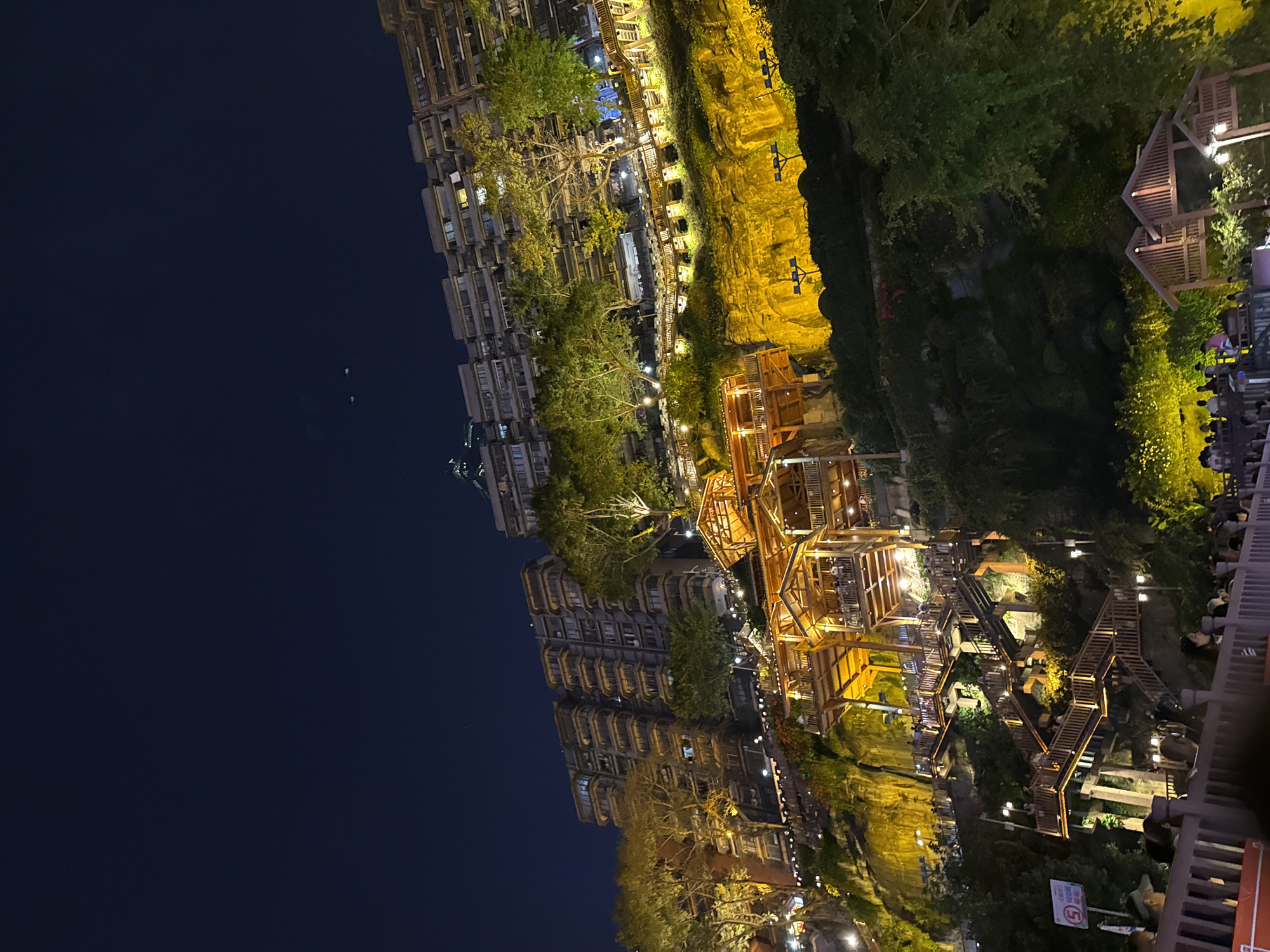 This nighttime image captures a vibrant, illuminated multi-level complex alongside a dark body of water, likely a river. On the left, towering multi-story buildings with numerous windows and balconies line the water's edge, partially obscured by trees. The water itself appears dark and still, reflecting a few faint lights.
The central and right portions of the scene showcase an elaborately lit terraced area. A prominent, rugged cliff face glows with warm yellow light, forming a dramatic backdrop. In front of and below this cliff are intricate wooden structures, including multi-tiered walkways, staircases, and platforms that appear to house outdoor dining or viewing areas. These structures are also warmly lit, creating an inviting ambiance. Lush green foliage and trees are interspersed throughout this illuminated landscape.
Numerous people are visible in the lower right, gathered at tables on the wooden platforms, suggesting dining or social activities. Others can be seen walking along the pathways and stairs. The activity appears to be leisure and entertainment, with people enjoying the scenic night view.
The time of day is clearly night, evidenced by the dark sky and extensive artificial lighting. The warm, golden hues of the lights highlight the architectural details and natural elements. The multi-level, terraced design, coupled with the tall buildings and river, strongly suggests a location like Hongyadong in Chongqing, known for its unique stilt-house architecture built into a hillside overlooking the Jialing River.
A white sign with red text is partially visible in the bottom right, displaying "COUNTY" and a circled number "5," possibly indicating a road or pedestrian sign.