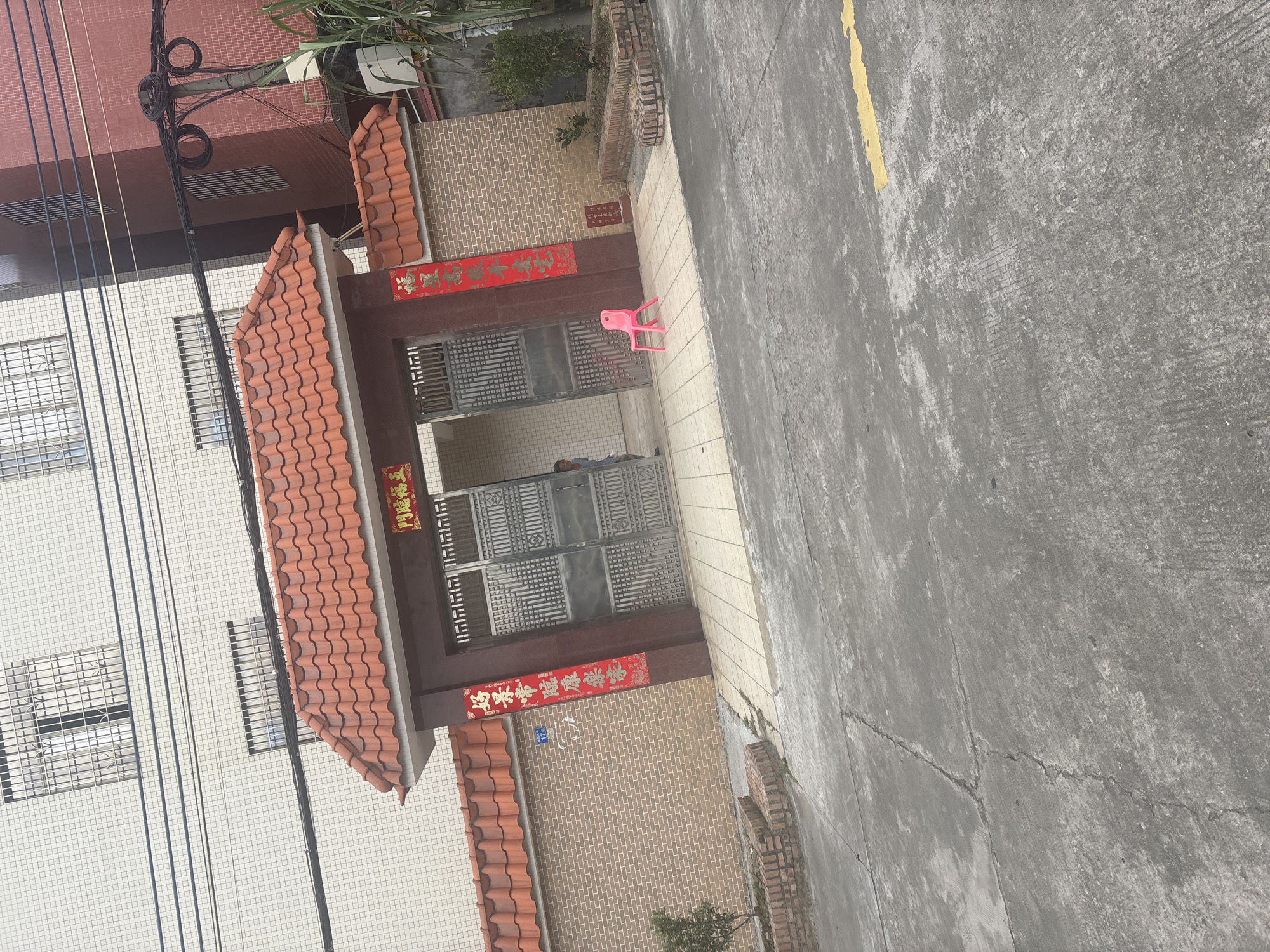 A weathered concrete ground leads to a decorative brick and concrete entryway with a tiled red roof, located in the Nanhai District, China. Two red banners with gold Chinese characters hang from the archway. The left banner reads "好孕带来健康平安" and the right banner reads "福". A person, possibly a child, is partially visible standing inside the doorway. A bright pink plastic chair sits on the tiled area in front of the entrance. A small tree is planted in a brick planter to the left, and a larger tree with green foliage grows to the right. The background features multi-story buildings with white and red brick exteriors, adorned with barred windows and electrical wires. A yellow painted line is visible on the concrete ground.