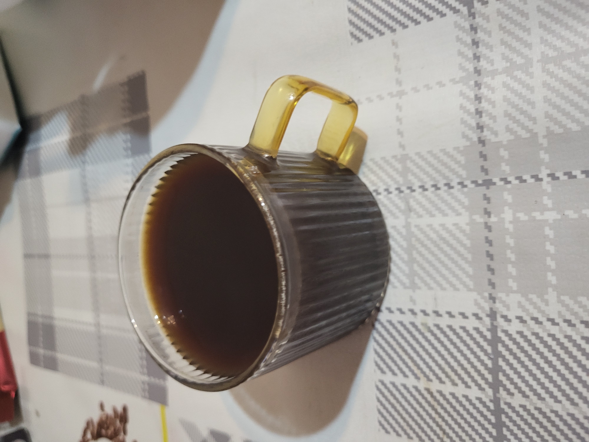 A clear, ribbed glass mug with a prominent amber-colored handle is depicted, containing a dark brown liquid, likely coffee or tea. The mug rests on a light-colored surface, which appears to be a tablecloth or placemat, adorned with a grey and white plaid pattern. The pattern features varying shades of grey in a grid-like design. The scene is set indoors, possibly at a home or cafe in Yerevan, Armenia. The image is oriented vertically, causing the mug to appear as if it's on its side. Reflections on the mug and the liquid indicate the presence of an indoor light source. No people or text are visible.