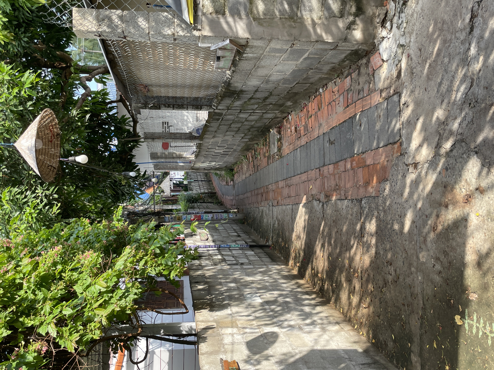 The image shows a narrow alleyway in Mũi Né, Vietnam. The alley is paved with a combination of concrete and red bricks, with a concrete walkway on the left and a brick and concrete path on the right. The walls on either side of the alley are made of grey concrete blocks. Lush green trees with some pink flowers hang over the alley from the left side, casting dappled shadows on the ground. A conical straw lamp with a lightbulb hangs from a tree branch over the alley. The alleyway curves into the distance, with buildings visible on both sides. A chain-link fence runs along the top of the wall on the right. There are no people or animals visible in the image. The overall impression is of a quiet, residential alley in a tropical climate.