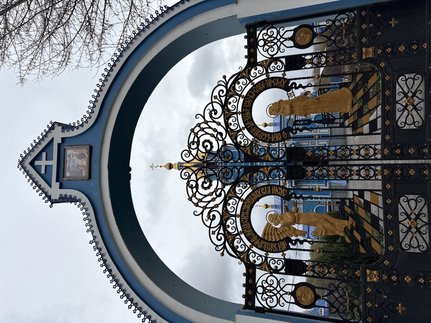 An ornate entrance gate and archway, likely belonging to an Orthodox church or monastery, are captured under an overcast sky. The image is oriented vertically, showing the structure from a slightly upward angle.
The archway, painted in striking blue and white, features intricate trim along its edge. At its apex, a white cross is set within a blue diamond-shaped element, beneath which a small, framed religious icon depicting several figures is embedded. Bare tree branches are visible against the cloudy sky to the left of the arch.
The large gate, appearing to be closed, is crafted from black wrought iron with extensive gilded ornamentation. It features intricate scrollwork, floral patterns, and geometric designs. Two prominent circular medallions are embedded within the gate, each containing a golden, three-dimensional religious figure. The upper medallion depicts a standing female saint, possibly the Virgin Mary, with a halo. The lower medallion shows a winged angel, likely Saint John the Baptist, with a halo. Around these figures, text in Old Church Slavonic or Cyrillic script is inscribed. The text around the upper figure partially reads "АГ-ІА СЛАВА ТРОІЧУ" (Holy Glory to the Trinity) and "МАТИ" (Mother). The text around the lower figure includes "Г-РА БЛАГОСЛОВЕН ІСУСЪ" (Lord, blessed Jesus) and "ІОАНЪ" (John).
Through the elaborate gate, parts of a church building are visible. It features a blue and white facade with architectural details and multiple domes, including gilded cupolas, characteristic of Orthodox Christian architecture. Green foliage can be seen near the base of the gate, hinting at landscaping within the grounds. The cloudy weather indicates a diffused light, possibly early morning, late afternoon, or simply an overcast day. The architectural style, iconography, and Cyrillic text are consistent with an Orthodox Christian site in Ulyanovsk, Russia.