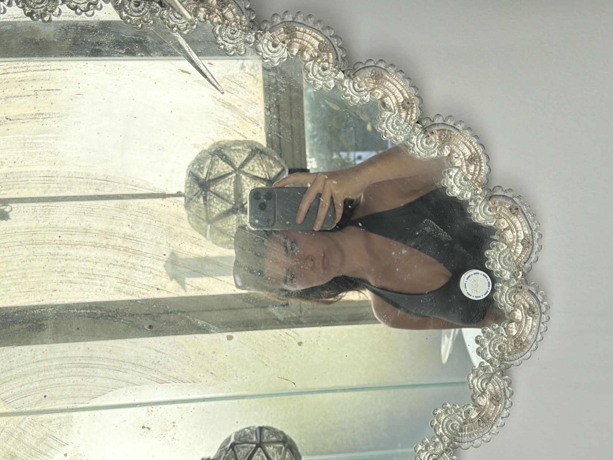 A person holds a smartphone in front of an ornate, antique mirror to take a selfie. The reflection shows the person’s face, shoulder, and arm in a black tank top, along with a second round, decorative object. The mirror is mounted indoors, with a window and light visible in the background. The photograph was taken in the city of Buenos Aires, Argentina.