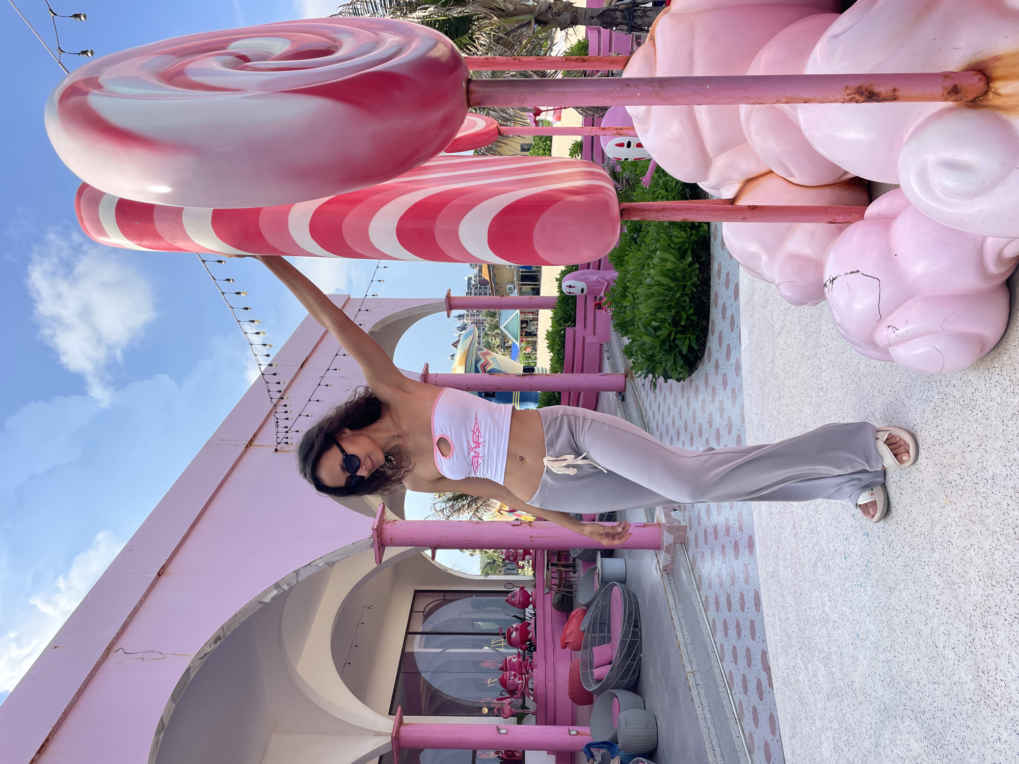 In Tien Thanh Ward, Vietnam, a woman stands in front of a pink building with arched entrances, leaning against a large, pink and white striped lollipop sculpture. She is wearing a white strapless top with a cutout and pink trim, and loose gray pants with a drawstring. Her left hand rests on the lollipop sculpture, and she is looking down with sunglasses on. To her left, several woven chairs and beanbag chairs are arranged on a patterned walkway. Behind the woman and to the right, more large pink candy-themed sculptures are visible, including a swirl lollipop and what appear to be marshmallow shapes. The background shows lush green foliage, palm trees, and parts of buildings, suggesting a resort or themed entertainment area. The sky above is blue with scattered clouds.