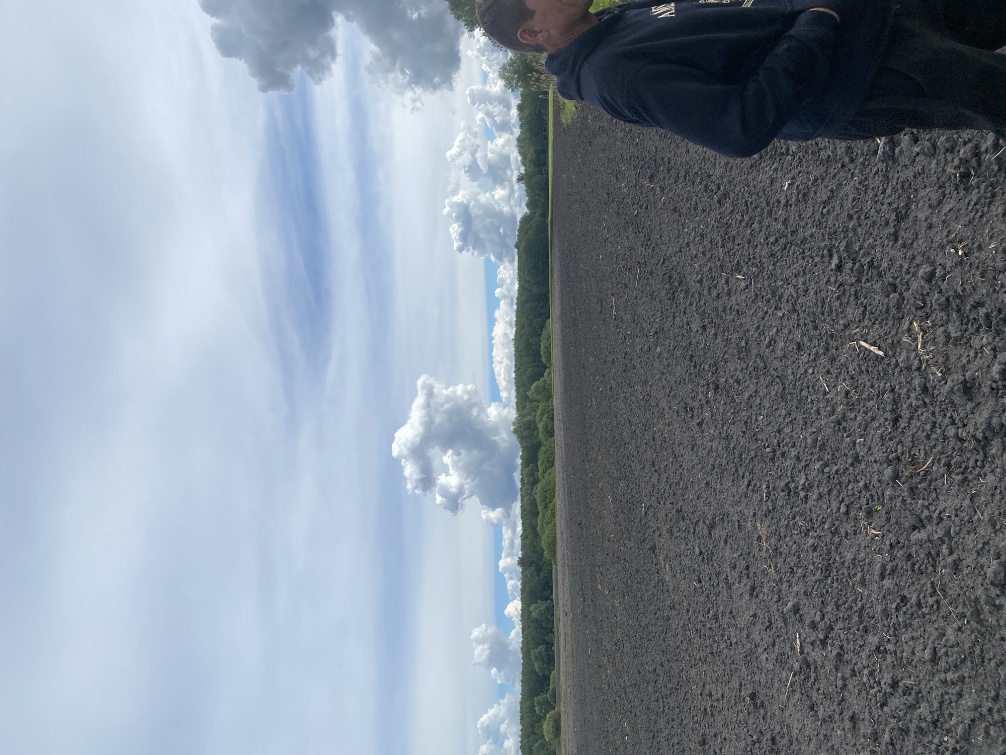 The image shows a landscape in Pidluky, Ukraine. A person stands on dark, tilled soil. A line of green trees separates the field from a sky filled with white and gray clouds. The sky transitions from cloudy to blue as it ascends in the image. The person is wearing a dark hoodie and pants. The view is slightly angled and the subject is at the right of the frame.