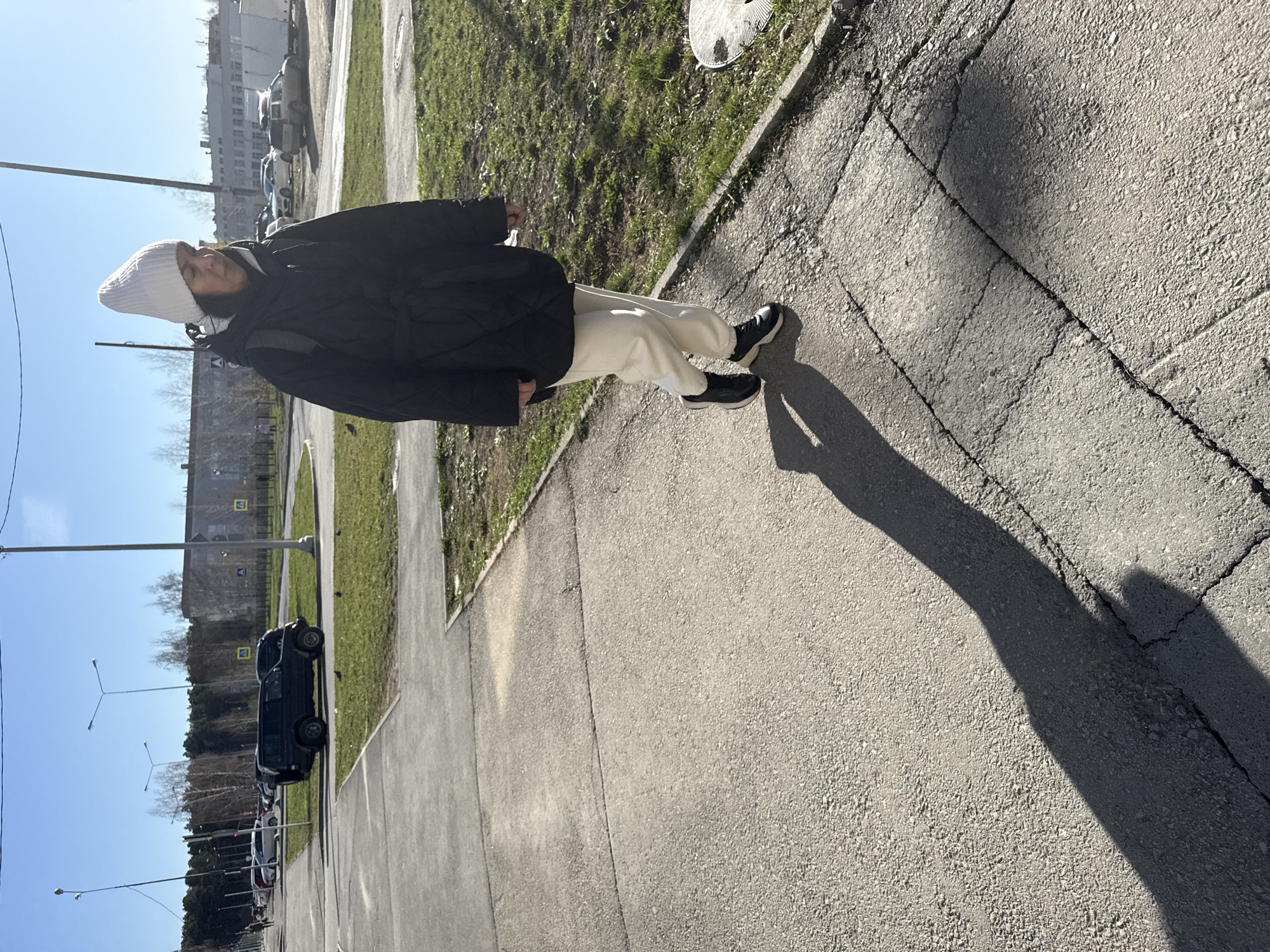 A full-body shot shows a person standing on a sidewalk in Yekaterinburg, Russia, on a sunny day. The person wears a white knit cap, a black winter coat, light-colored sweatpants, and black shoes. They cast a long shadow on the sidewalk. The sidewalk is concrete with cracks. A patch of green grass borders the sidewalk. Across the street, there are parked cars, trees, lampposts, and buildings. The sky is blue.