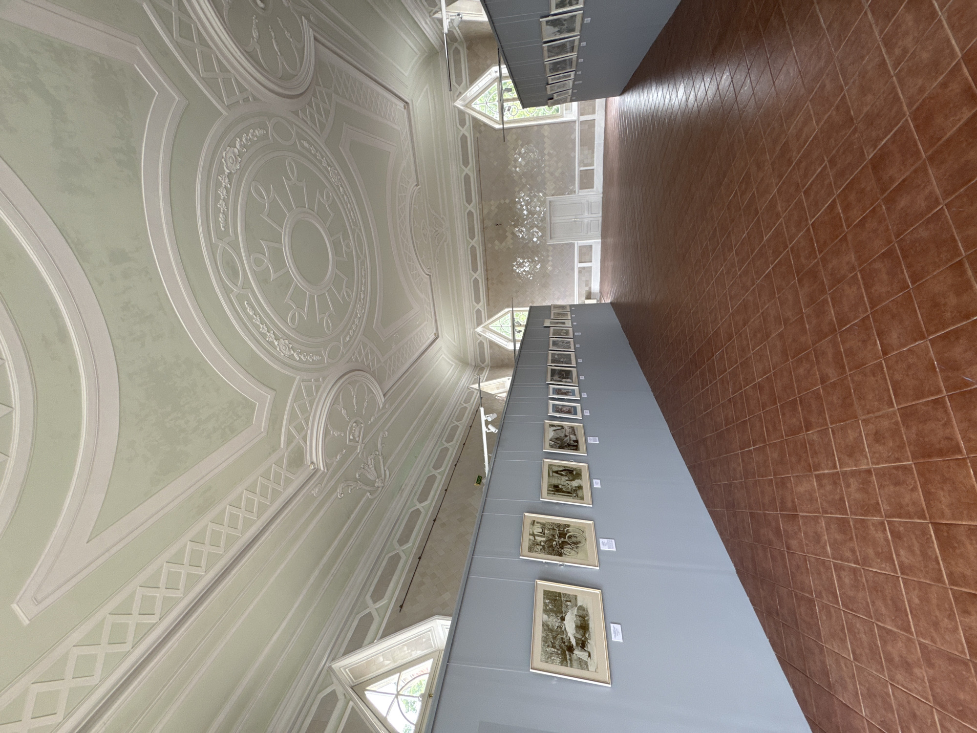 This is a photograph of the interior of an art gallery or exhibition space, likely located in Pushkin, Russia, given the ornate ceiling and architectural style reminiscent of historical estates in the area. The room features a high, decorative ceiling with intricate plasterwork in light green and white. The walls are a pale grey on the left and a textured, pale yellow with a diamond pattern on the back wall. A brown tiled floor with a diamond pattern covers the ground.
On the left wall, a temporary exhibition partition displays several framed artworks, which appear to be historical prints or paintings depicting scenes of people and daily life. These frames are light-colored. Further down the left wall, more artworks are visible. On the right, another partition displays more framed artworks, extending into the distance.
The room is spacious and empty of people, suggesting a quiet and unoccupied moment within the gallery. Natural light streams in from arched windows at the far end of the room, and the space is illuminated by artificial lighting tracks suspended from the ceiling. In the distance, double doors are visible on the back wall. The overall impression is one of a well-maintained exhibition space within a grand historical building.