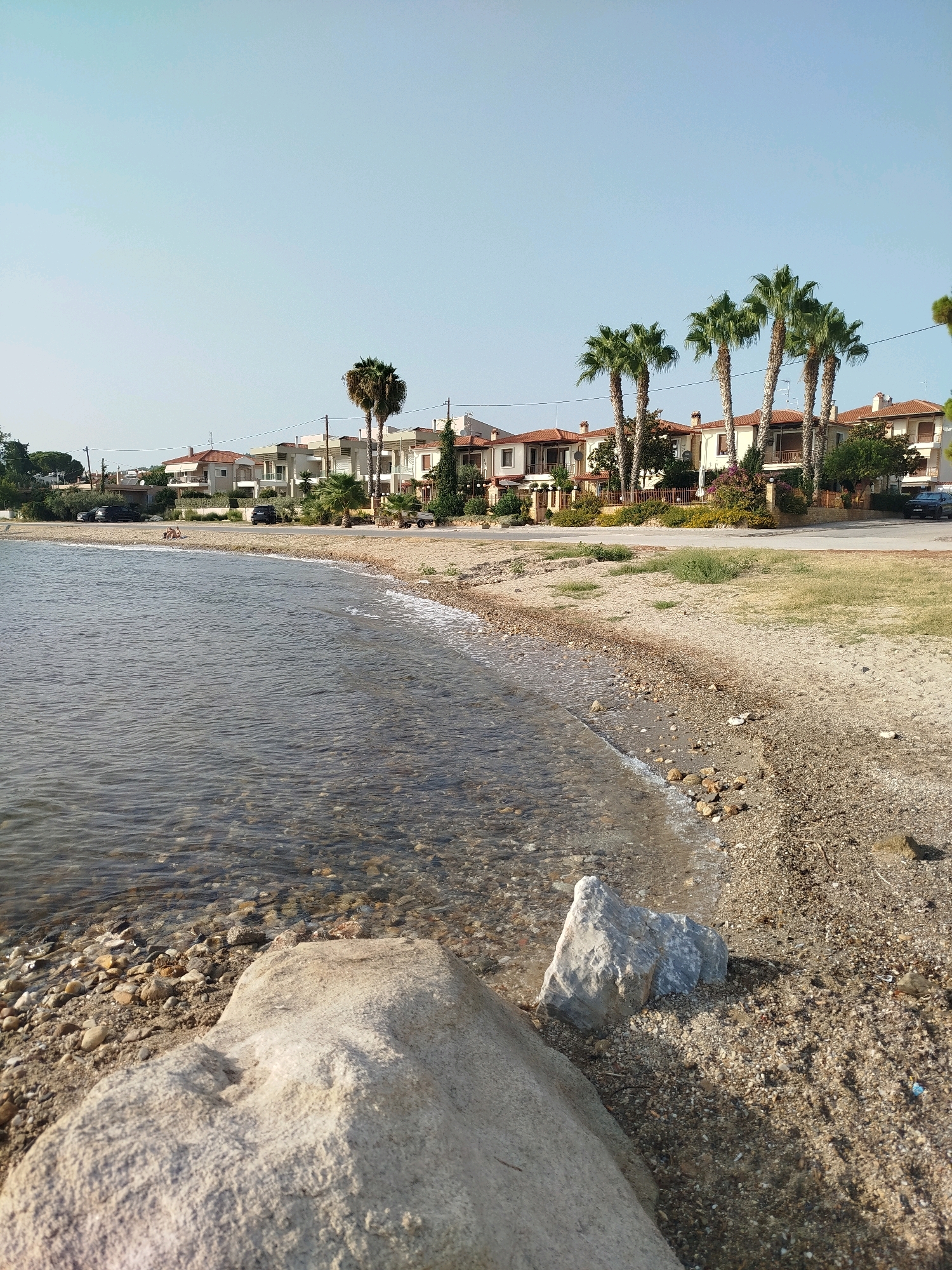 The image depicts a coastal scene in the Sithonia Municipal Unit, Greece. In the foreground, a rocky beach meets the clear, shallow water of the sea, where pebbles and small stones are visible beneath the surface. A large, light-colored boulder occupies the lower left portion of the frame. Beyond the beach, a row of modern houses with red-tiled roofs lines a street. Several tall palm trees are interspersed with the houses. A few cars are parked along the street, and two individuals are seated on the sandy area between the beach and the street. The sky is clear and blue, indicating a sunny day. The overall impression is of a quiet residential area adjacent to the coast.