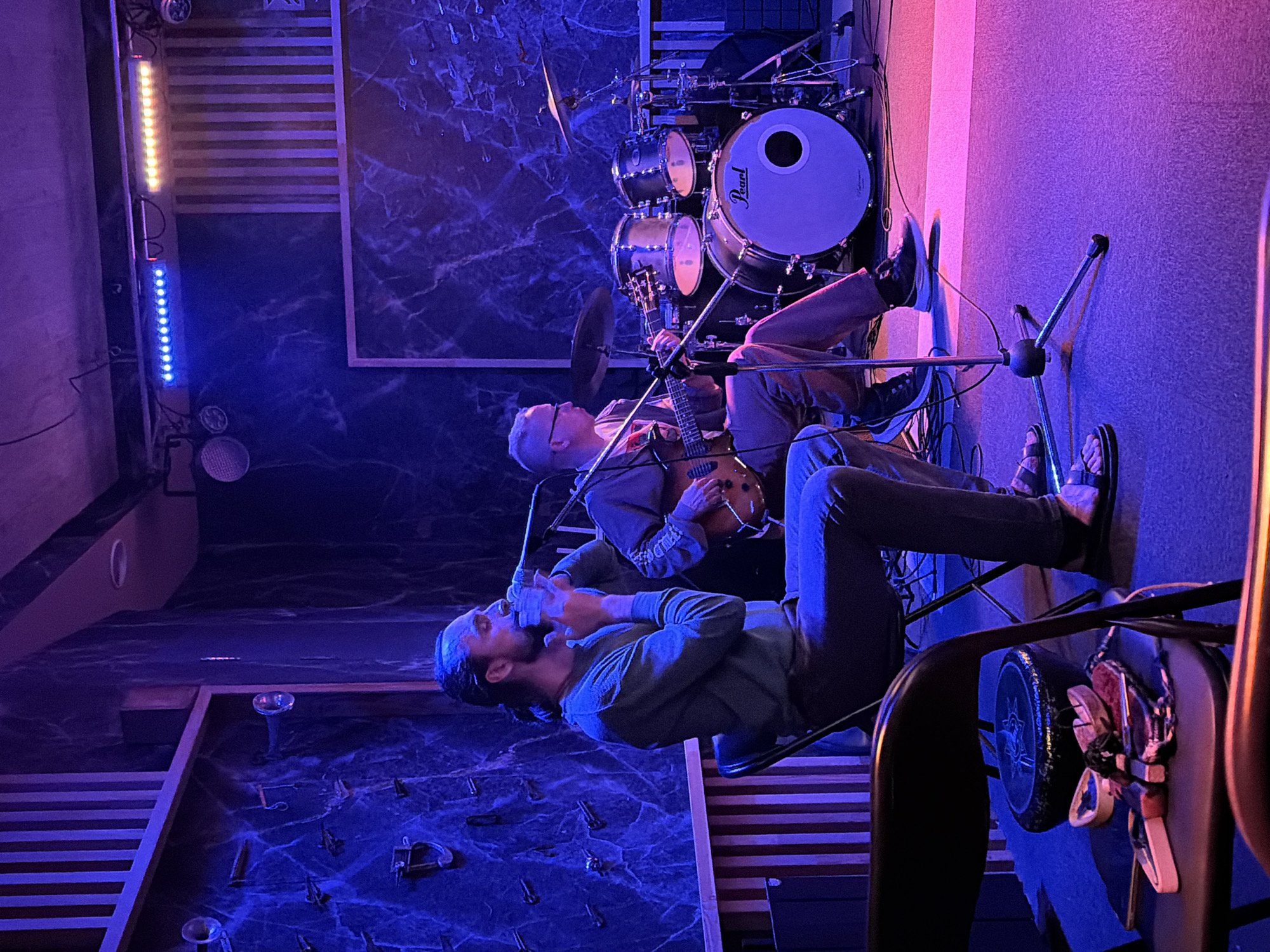 The image depicts an indoor scene with two individuals engaged in a musical performance. One person, seated, is playing an electric guitar and appears to be singing into a microphone. This individual wears glasses and a grey jacket. The other person, also seated, is singing into a microphone. They have a beard and are wearing a long-sleeved green shirt.
Behind the guitarist, a drum set is visible, suggesting a band performance. The setting is dimly lit, with vibrant blue and purple lighting casting dramatic shadows. The walls have a dark, marbled texture. Striped surfaces, possibly a wall or stage element, are also present in the background.
The overall atmosphere is that of a live music performance, possibly in a small venue or studio. The lighting suggests it's nighttime or an intentionally dark environment. The focus on the musicians and their instruments indicates the central activity is the music. There is no visible text in the image.