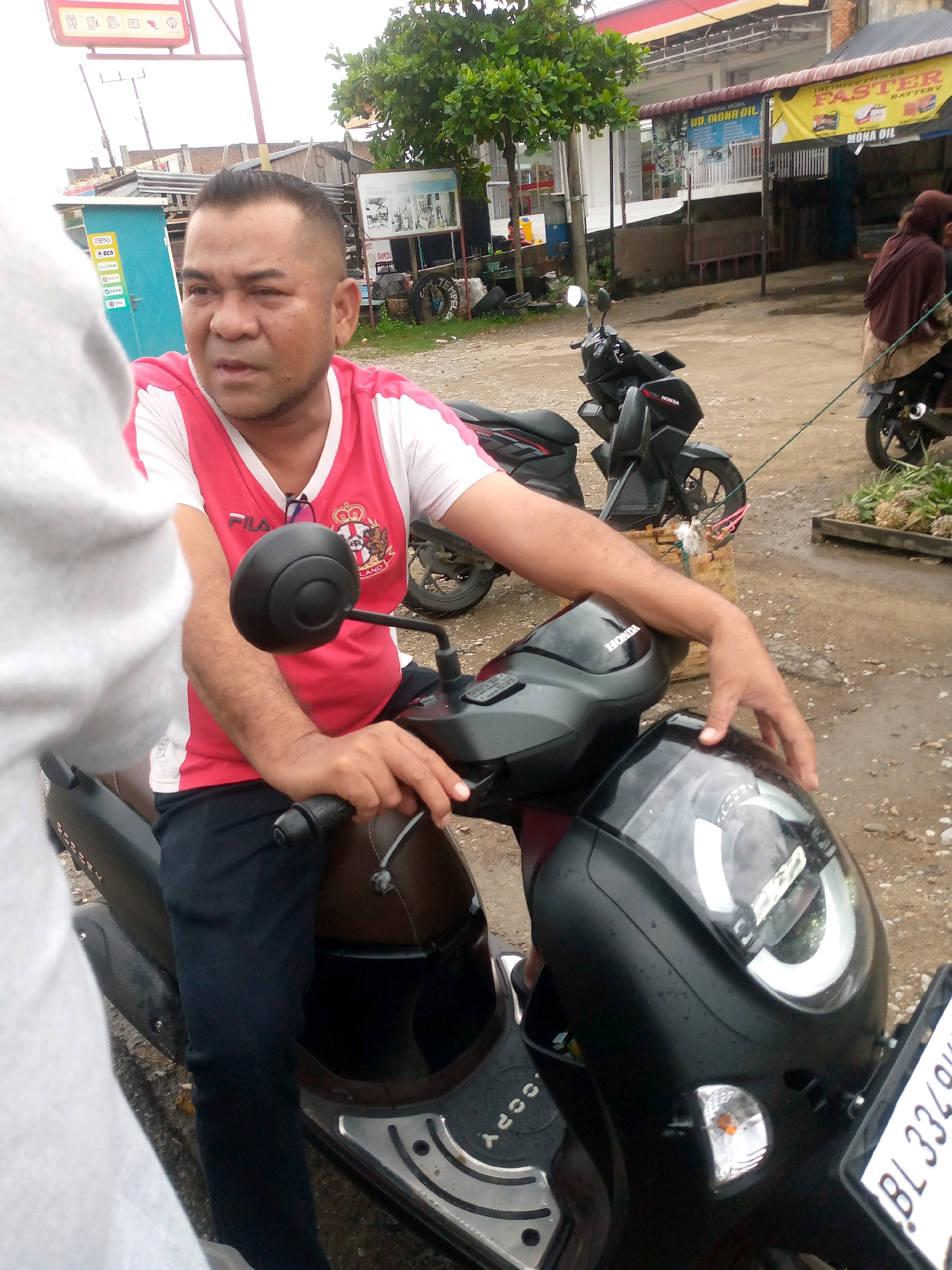 A man sits on a dark-colored Honda scooter parked on a gravel-covered road. The man is wearing a red and white v-neck shirt and dark pants. He has a short haircut and appears to be looking to his left. His left hand is on the scooter's handlebar. The scooter has a black front fairing with a prominent headlight. A license plate with the characters "BL 3334 0" is visible on the front. In the background, there are buildings, trees, and other parked motorcycles. A yellow banner with text that includes "FASTER BATTERY" and "MONA OIL" is visible on a building. Another building has blue and white signage, including "UD. MONA OIL". The overall environment suggests a commercial area in a town or city. The weather appears to be overcast or recently rainy, as evidenced by water droplets on the scooter's fairing.