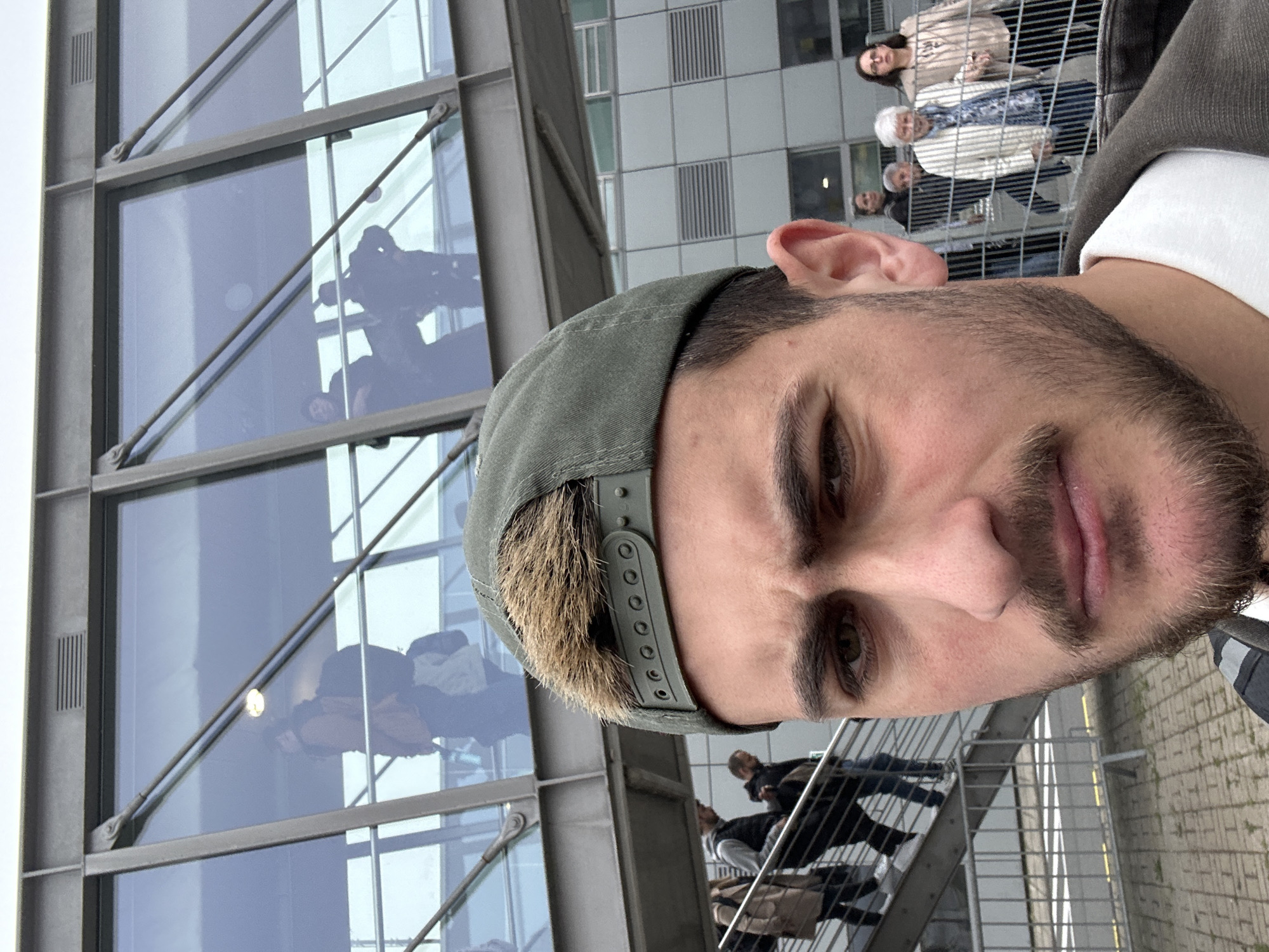 A tilted selfie of a man with a beard and a green baseball cap, taken near a modern building with large windows reflecting people. In the background, there are several people walking near a building. The scene is likely in Dortmund, Germany, as evidenced by the architectural style of the building.
