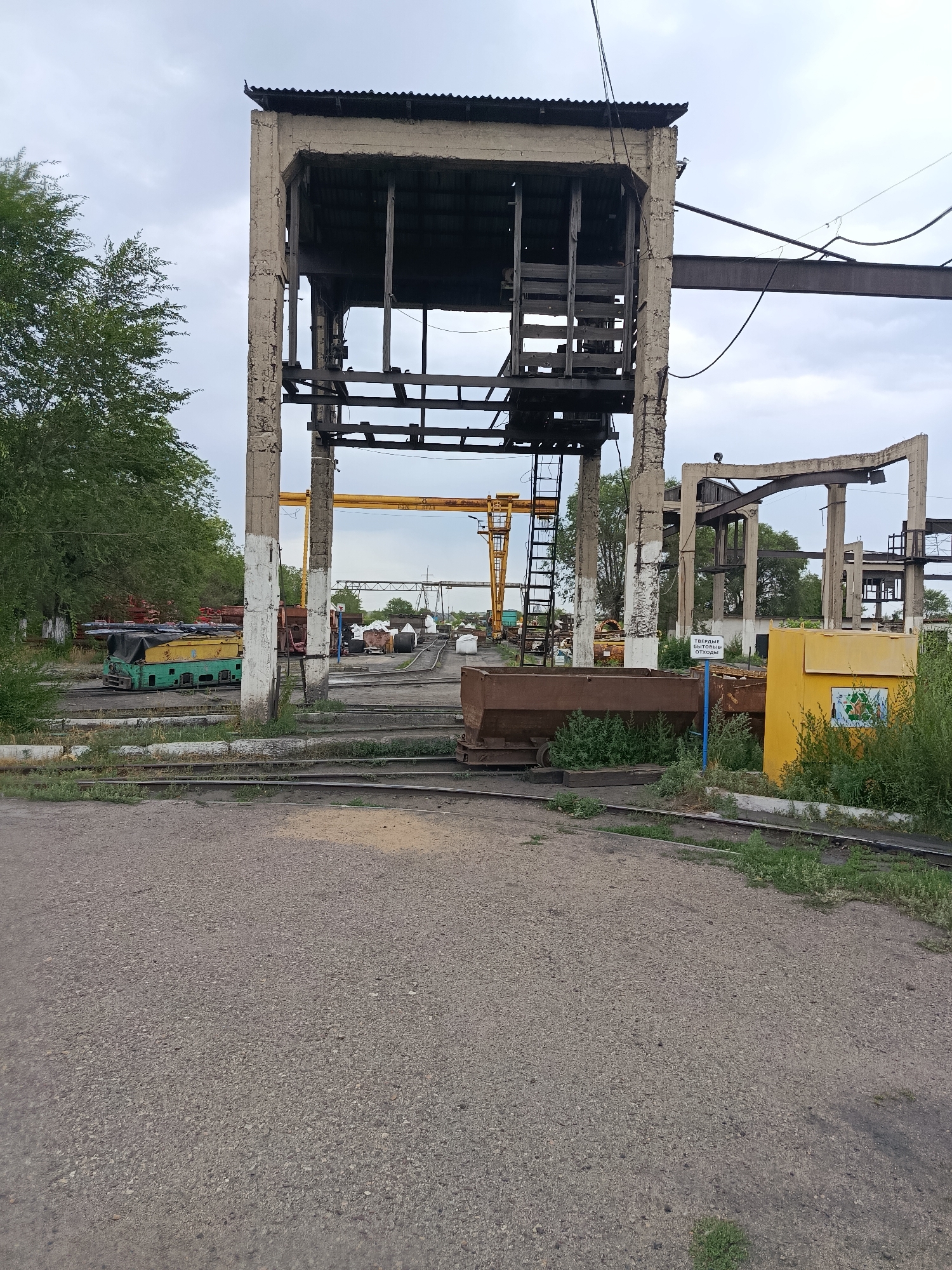 The image shows an outdoor industrial or junkyard scene under a cloudy sky, likely located in Saran, Kazakhstan. In the foreground, there is a gravelly, light-colored surface with several parallel railway tracks running through it. To the left, lush green trees are visible. Dominating the center of the image is a large, dilapidated concrete structure, appearing to be a former industrial building or loading dock, with exposed wooden beams and metal framing. A black ladder extends up the side of this structure. To the right of the concrete structure, a bright yellow bin with recycling symbols is positioned near more overgrowth. In the background, various industrial elements are present, including what appears to be a yellow gantry crane, other railway cars or containers, and a flat, overcast sky. The overall impression is one of disuse and decay within an industrial setting. There are no people visible in the scene.
